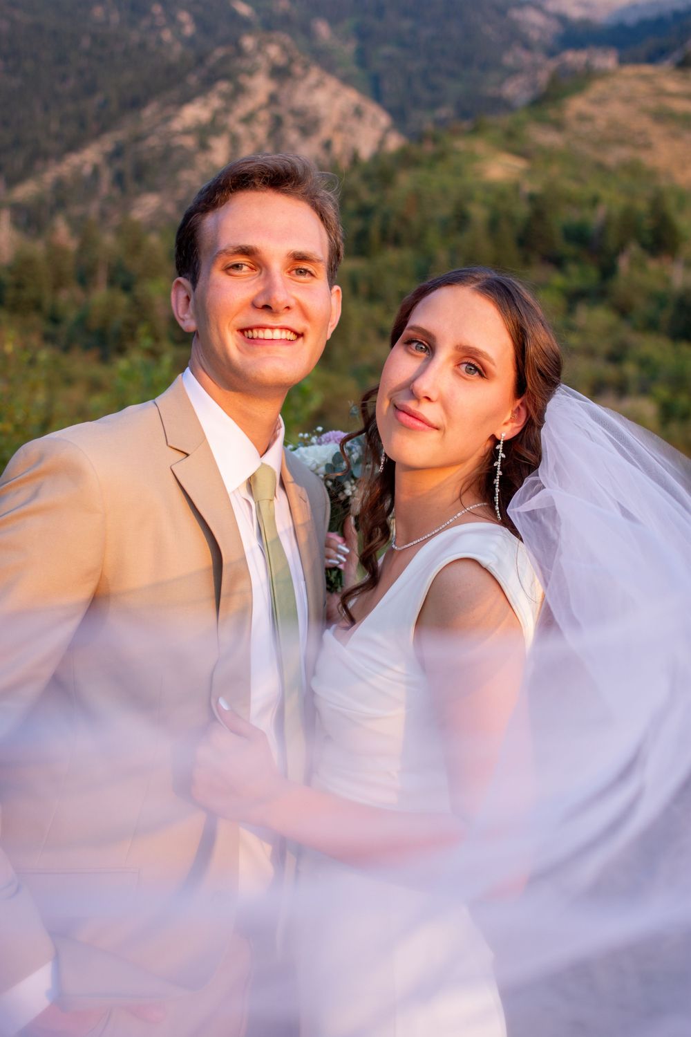 Bride and groom smile at camera as veil floats towards camera with an Utah mountain landscape in background.