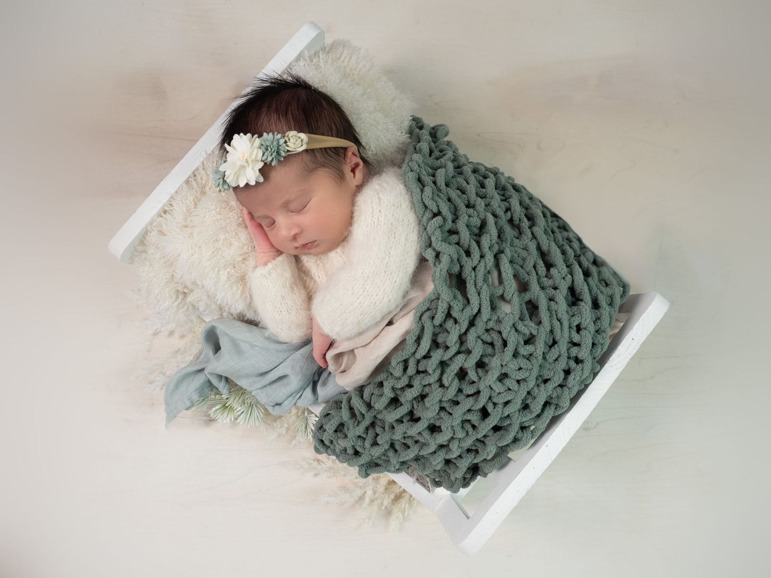 Newborn baby girl sleeps peacefully in a little white bed, wrapped in soft pastel taupe and green and white - a dreamy, gentle moment