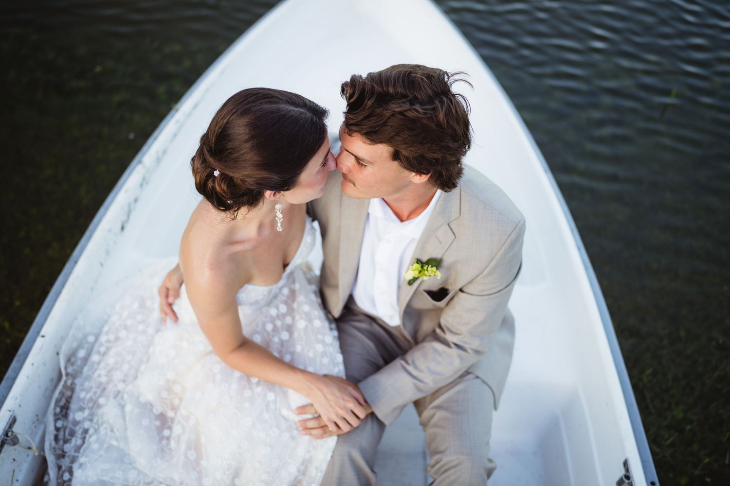 Newlywed couple shares intimate moment while sitting in white boat on dark water during wedding photoshoot.
