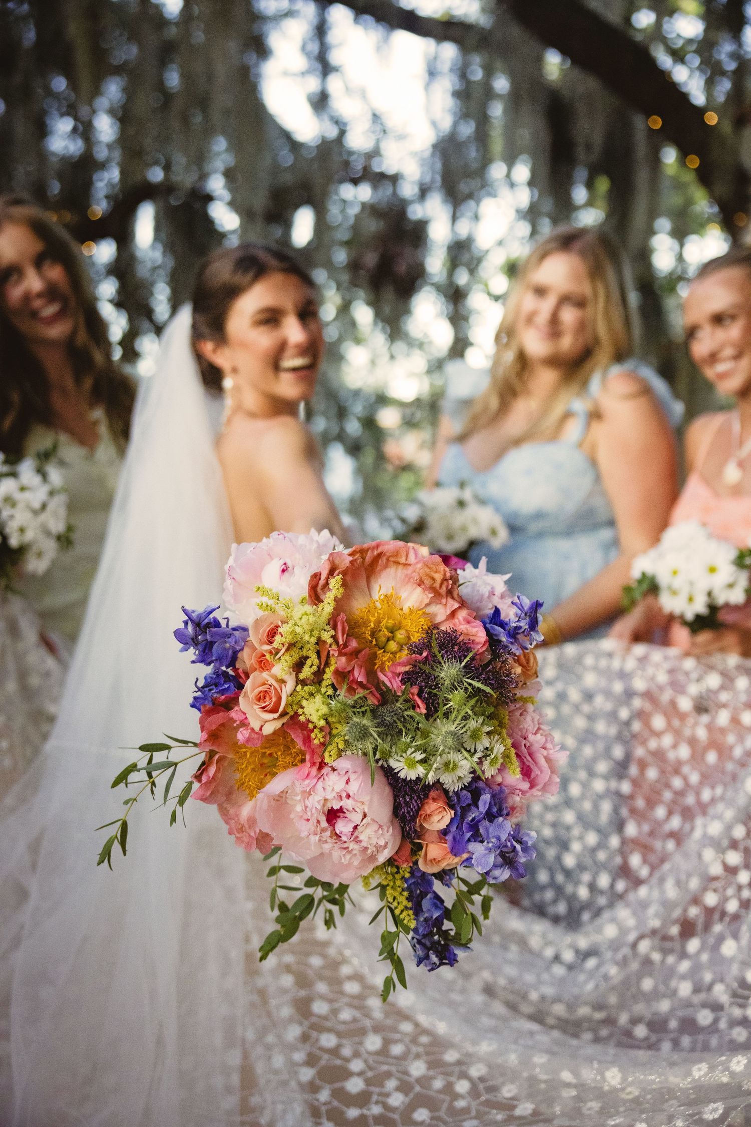 A vibrant wedding bouquet with pink peonies, purple delphinium, and orange ranunculus against blurred bridesmaids dresses.