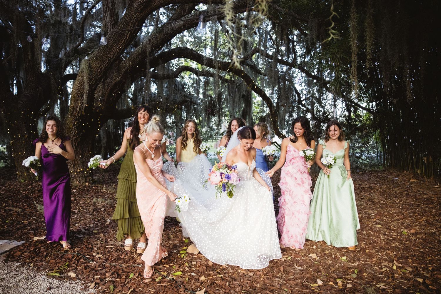 A bridal party in pastel dresses poses under large trees in a forest setting during an outdoor wedding celebration.