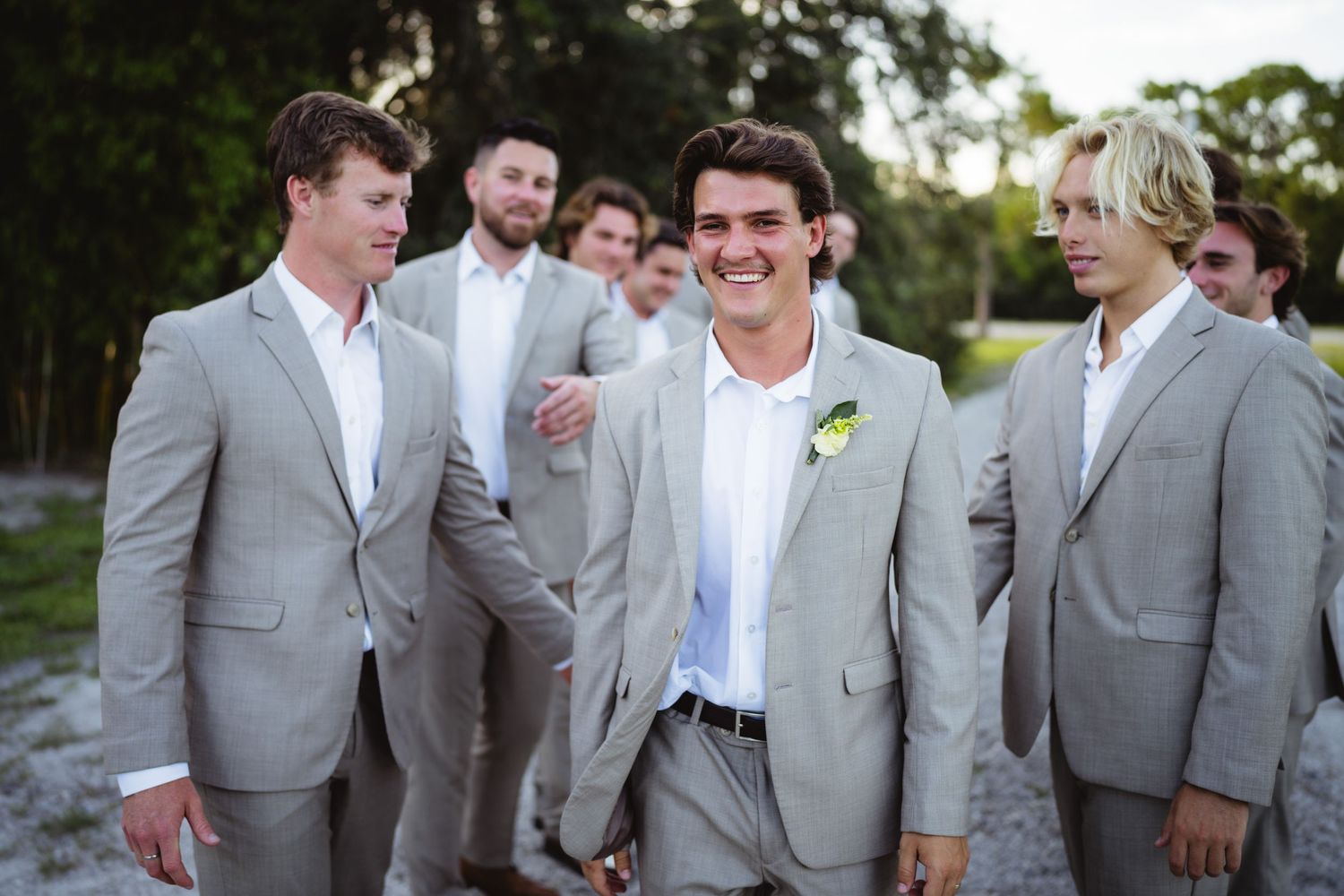 Groomsmen wearing light gray suits and white shirts smile together during outdoor wedding photographs.