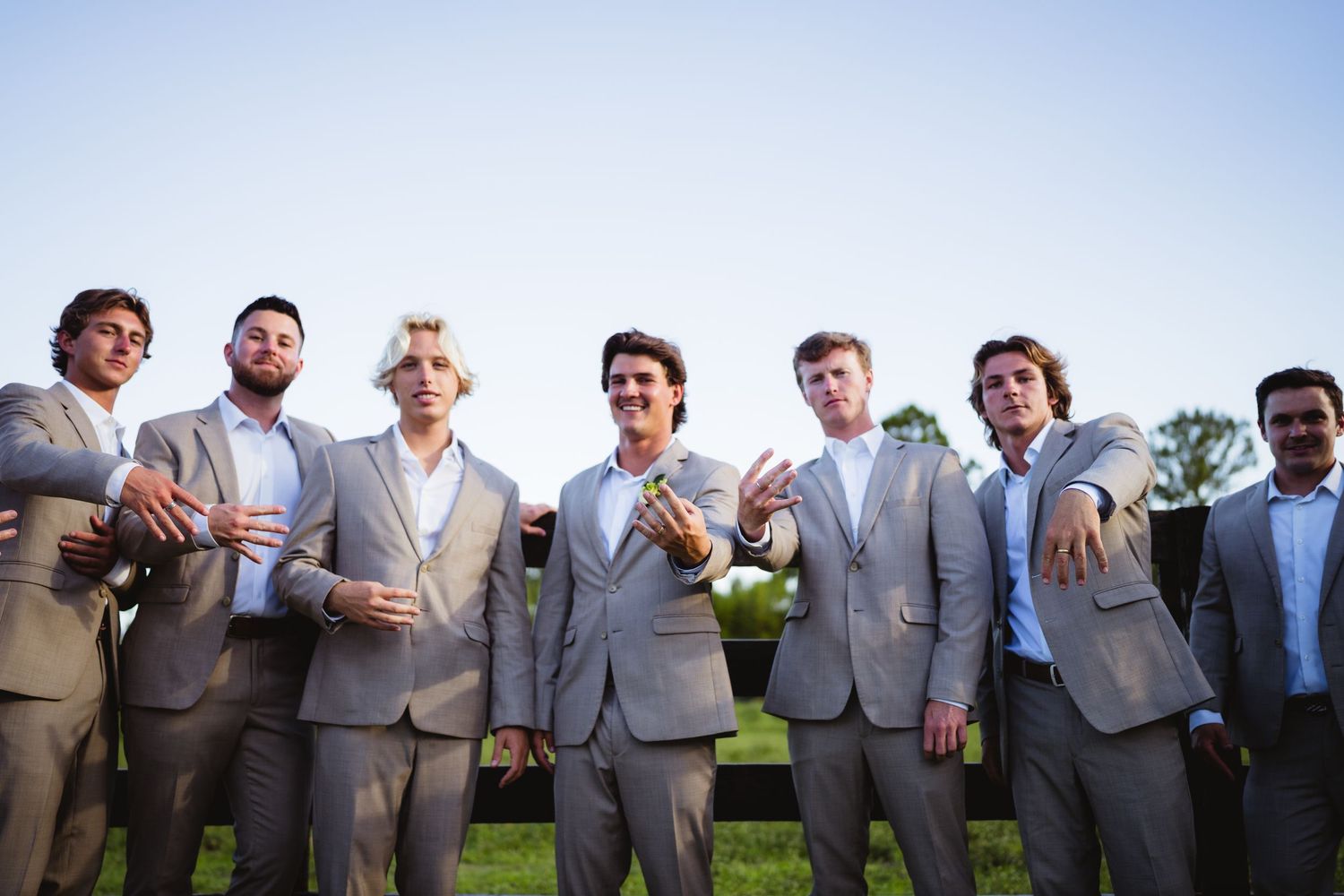 A group of smiling groomsmen wearing matching light gray suits pose together outdoors.