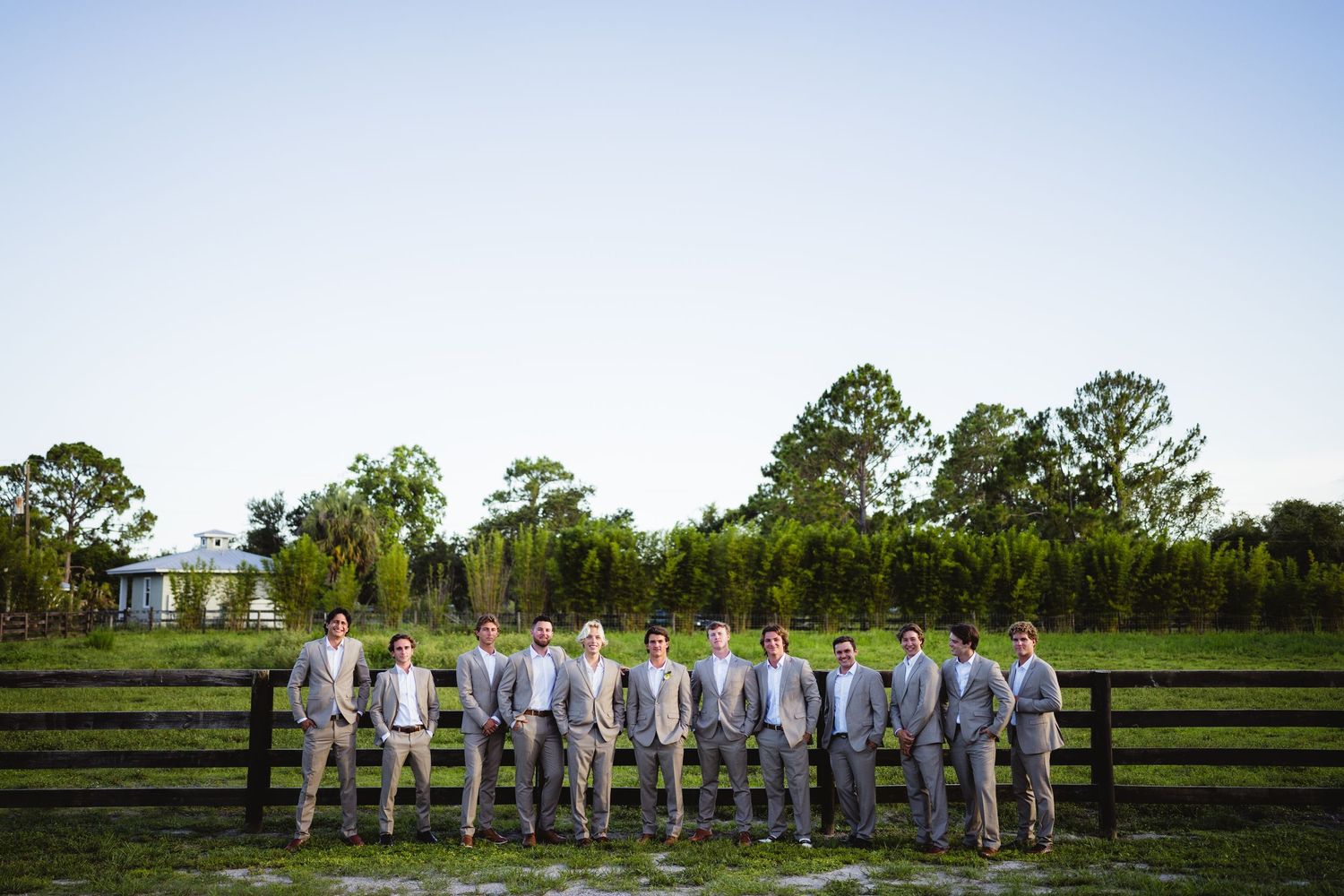 Groomsmen in light gray suits stand in a line against a wooden fence on a sunny day at an outdoor venue.