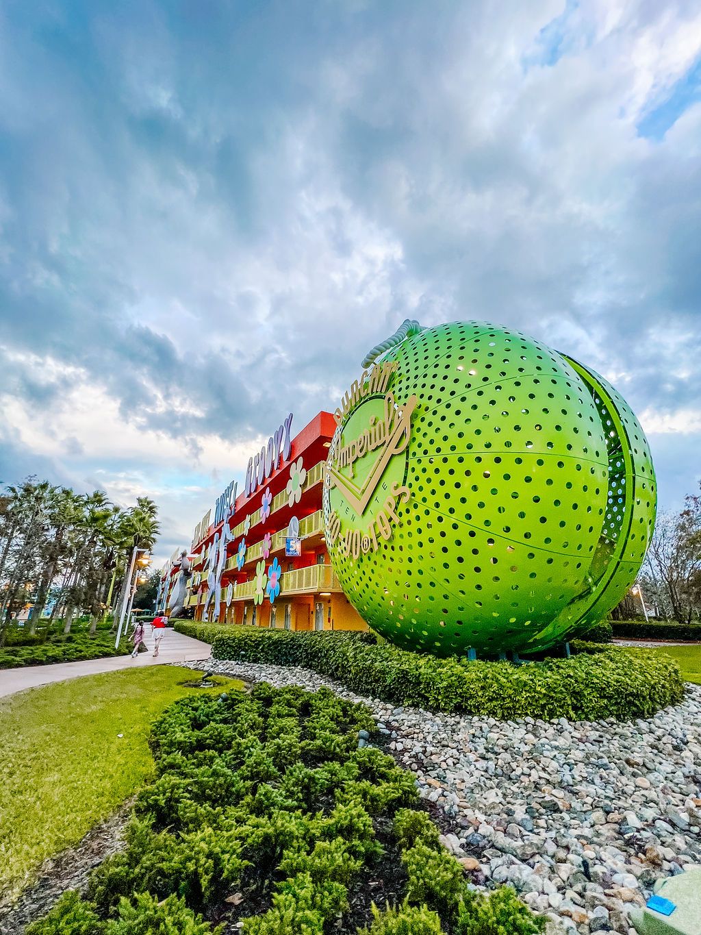 A giant green tennis ball sculpture sits in front of a modern colorful building with dramatic cloudy sky overhead.
