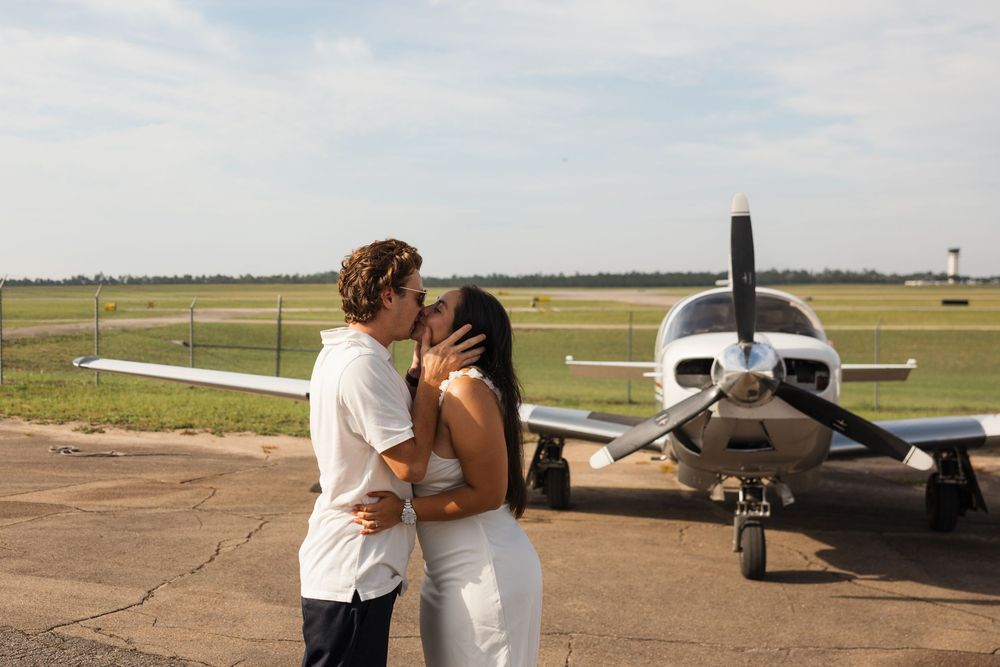 A small private aircraft parked on a sunny runway at a small airport with grass fields visible in the background.