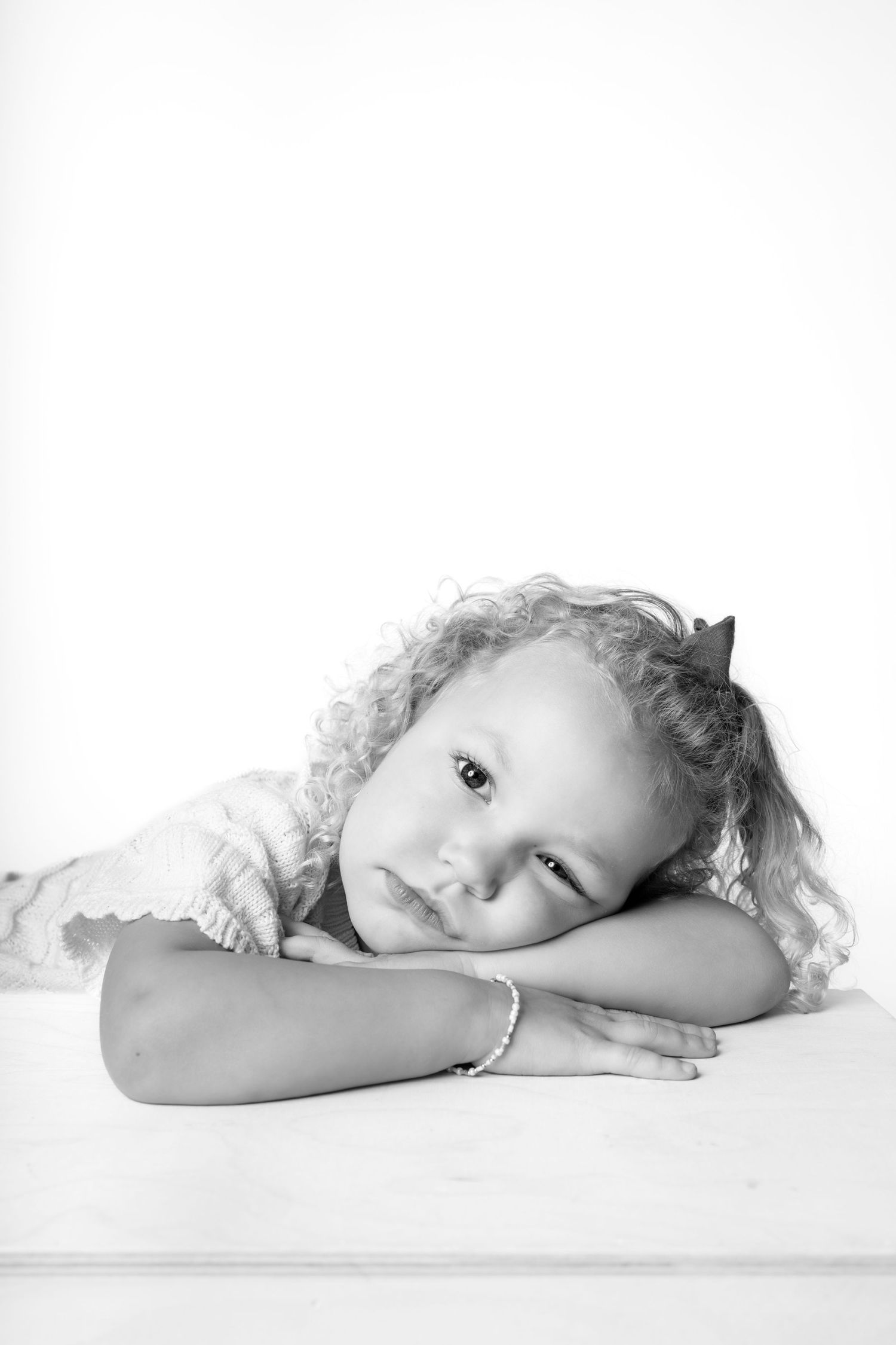 Black and white portrait of a young curly-haired girl resting her head on her arms on a white surface.