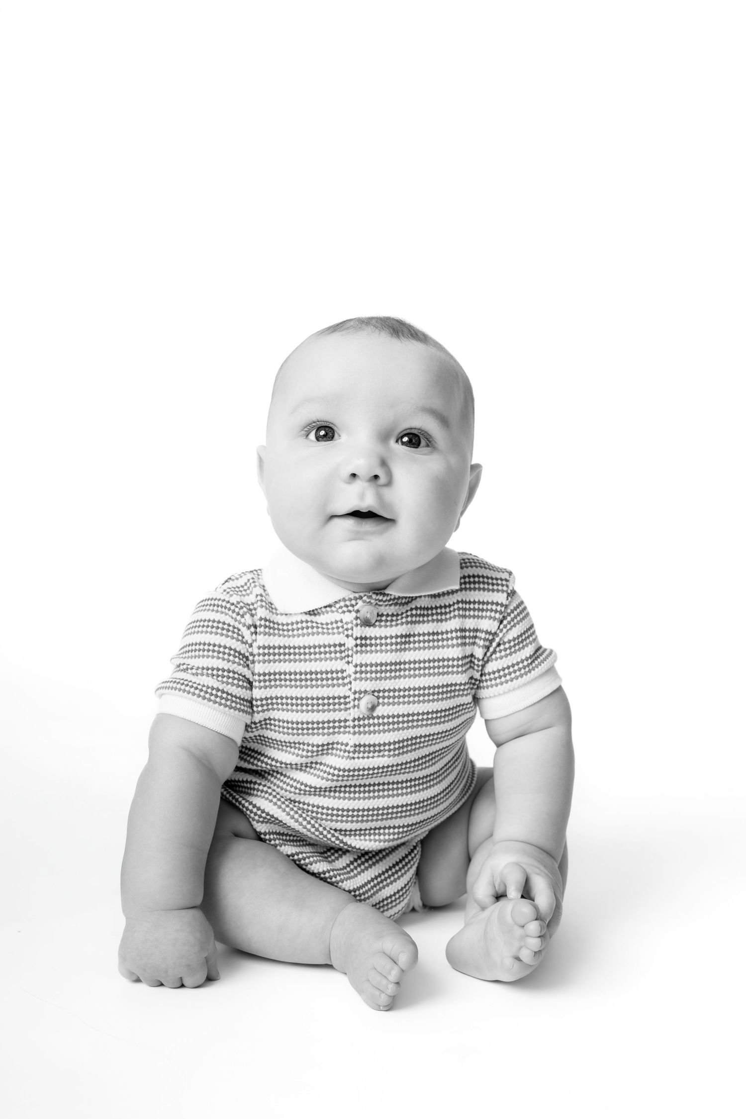 Black and white portrait of a cute baby sitting upright, wearing a striped onesie, looking up curiously.