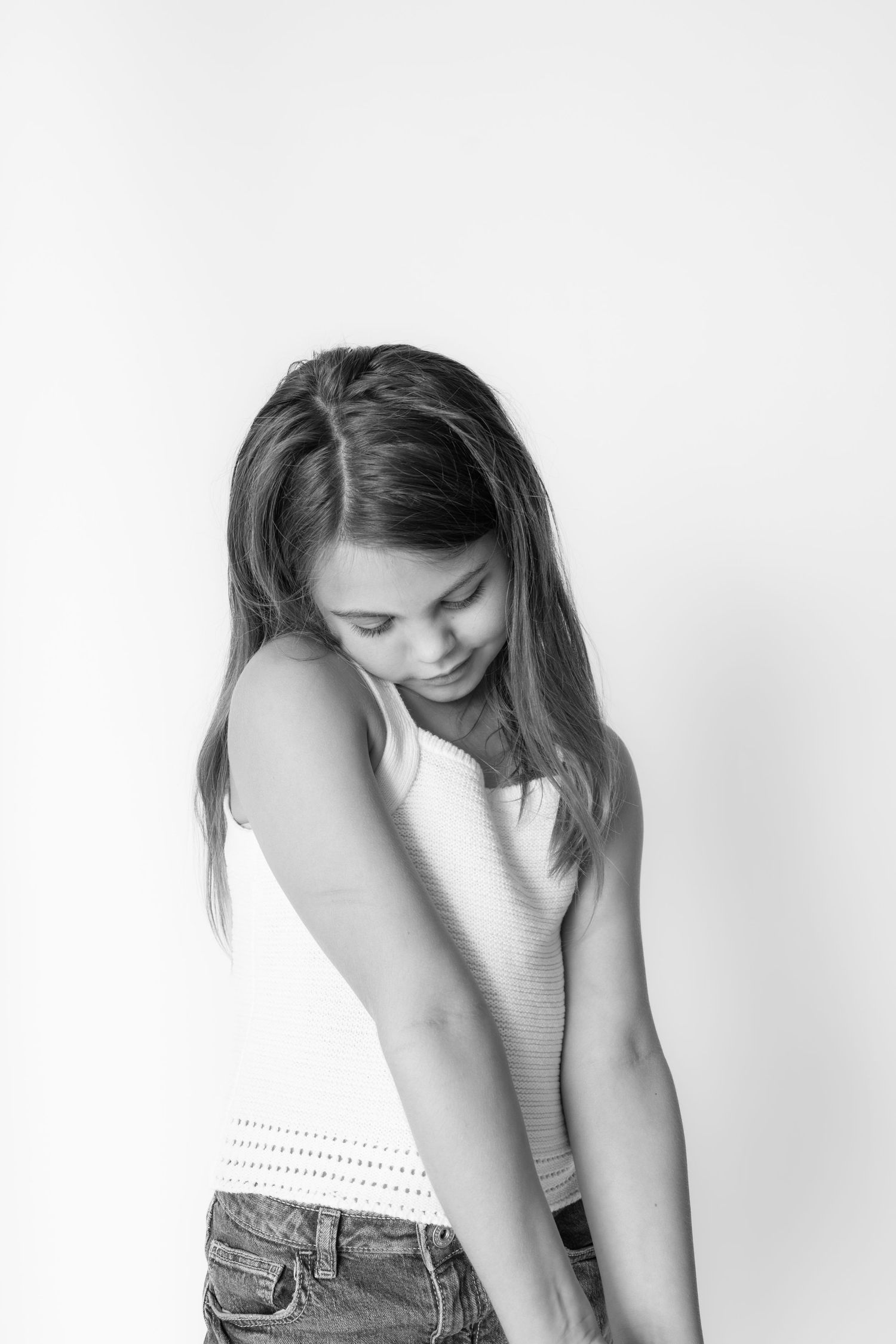 Black and white portrait of a young girl in a white tank top and jeans, looking down against a white background.
