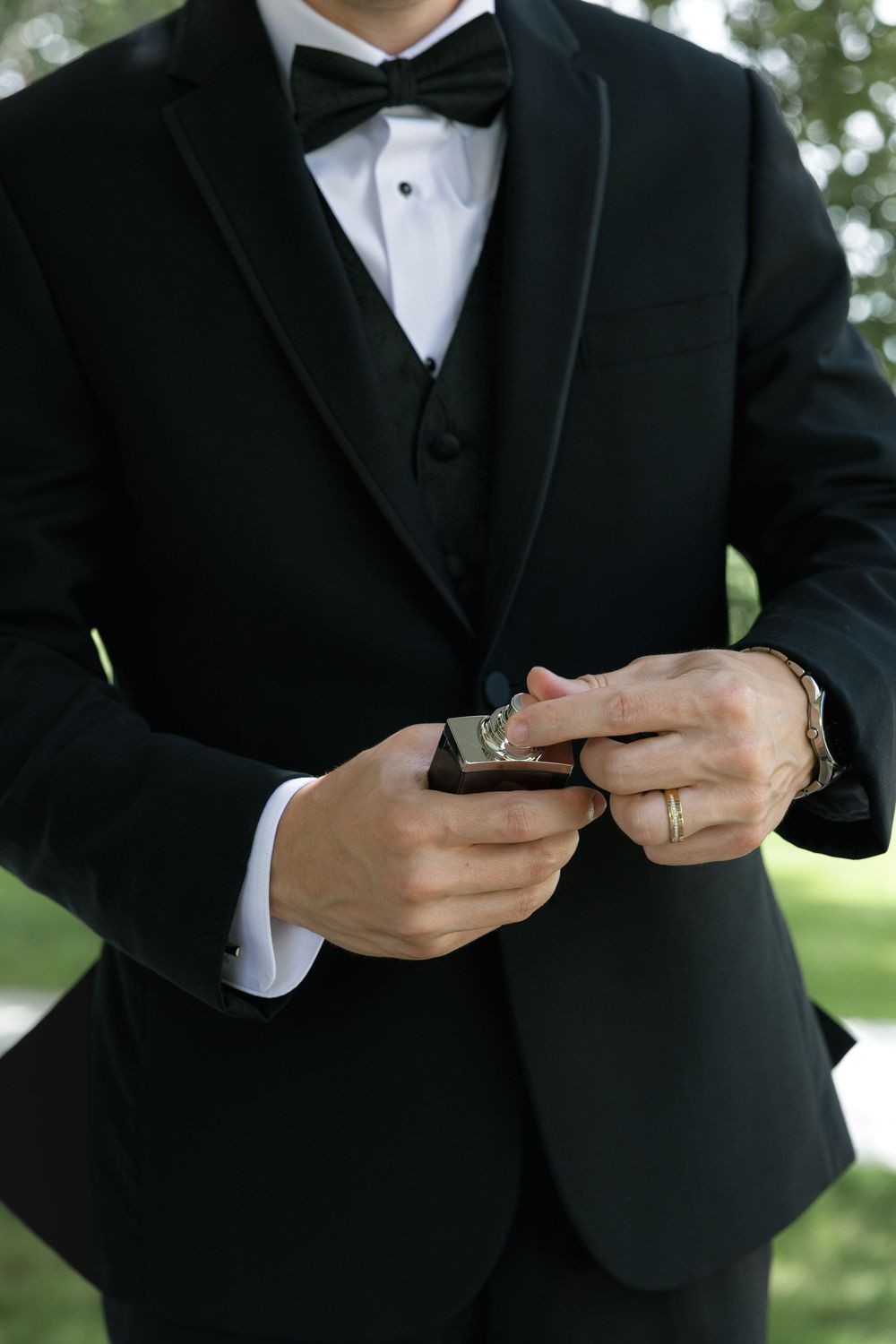 Close-up view of a groom in black tuxedo and bow tie adjusting cufflinks during a wedding ceremony outdoors.