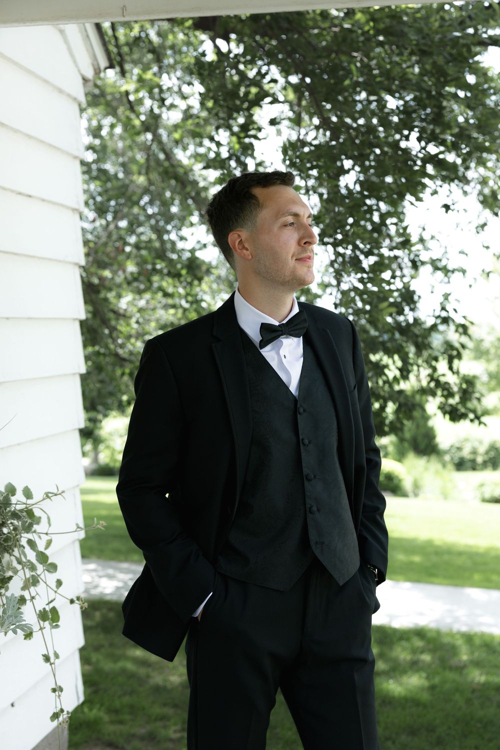 A groom in a classic black tuxedo and bow tie stands pensively near a white wall on a sunny outdoor day.