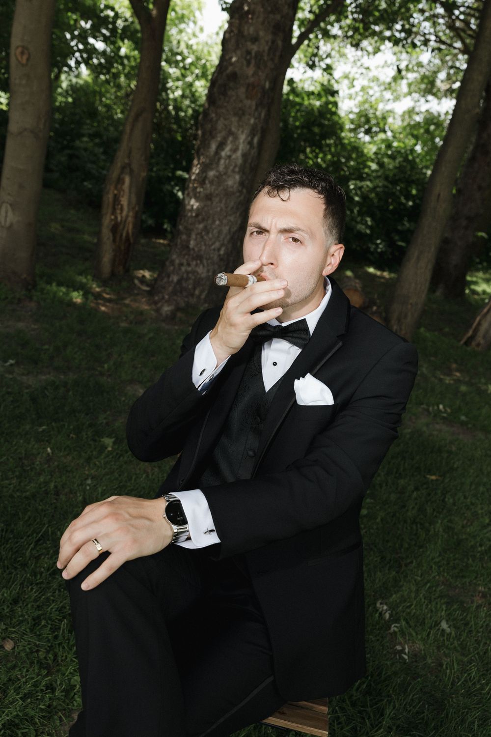 Groom in black tuxedo and bowtie sitting on bench in wooded park setting taking candid photos.