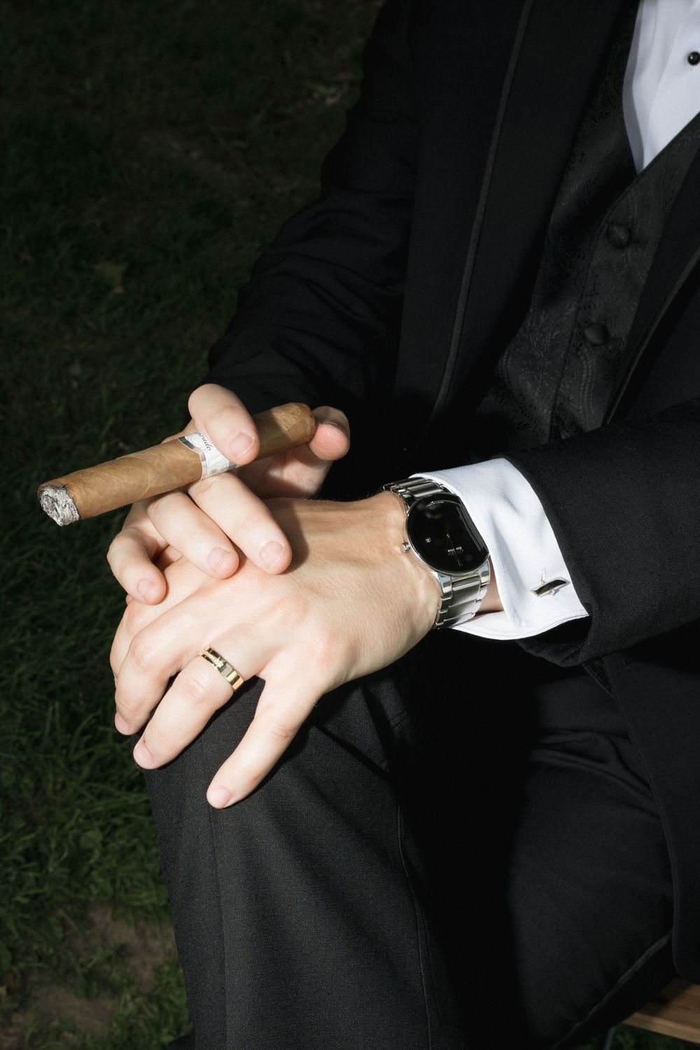 Close-up of hands holding a cigar, wearing a black suit and silver wristwatch with a wedding ring visible on the finger.