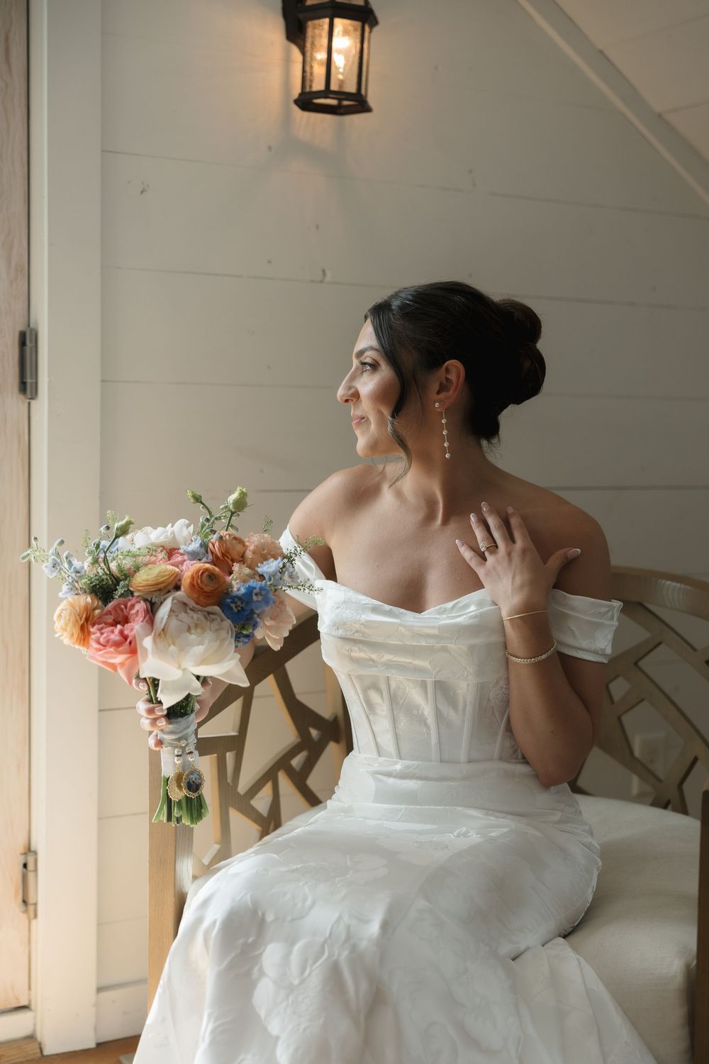 Elegant figure in white strapless wedding dress holds colorful bouquet while sitting in vintage chair by lantern-lit doorway.