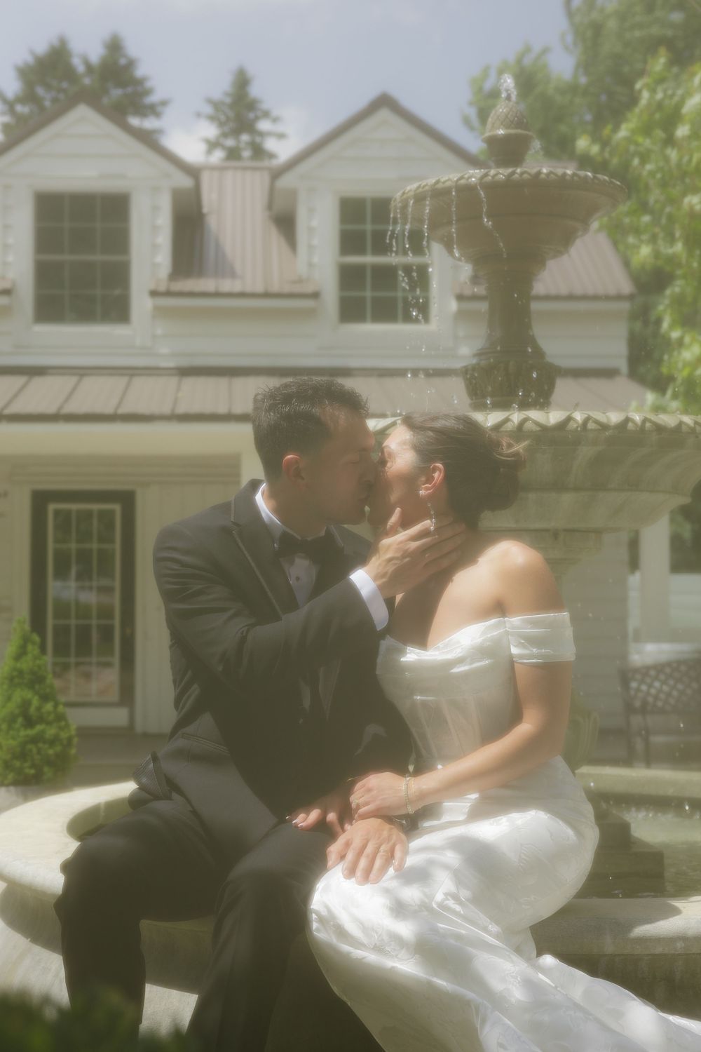 A newlywed couple shares a romantic kiss on a sunny day with white houses and trees in the background.
