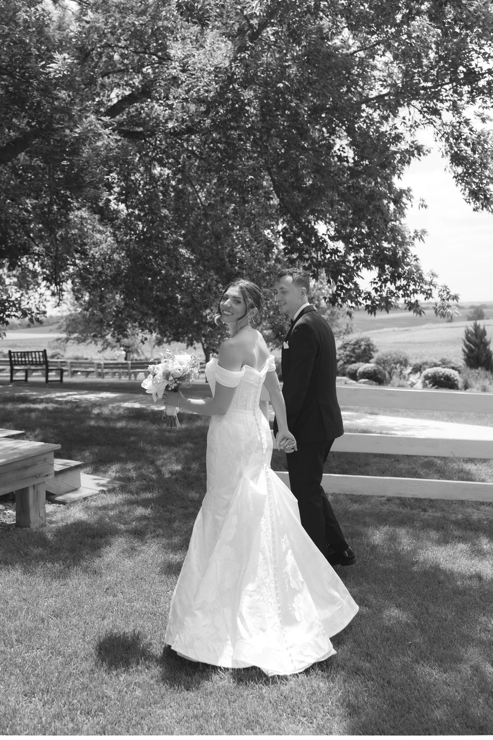 A couple dances together on grassy grounds with trees in the background, her white gown flowing elegantly in the movement.
