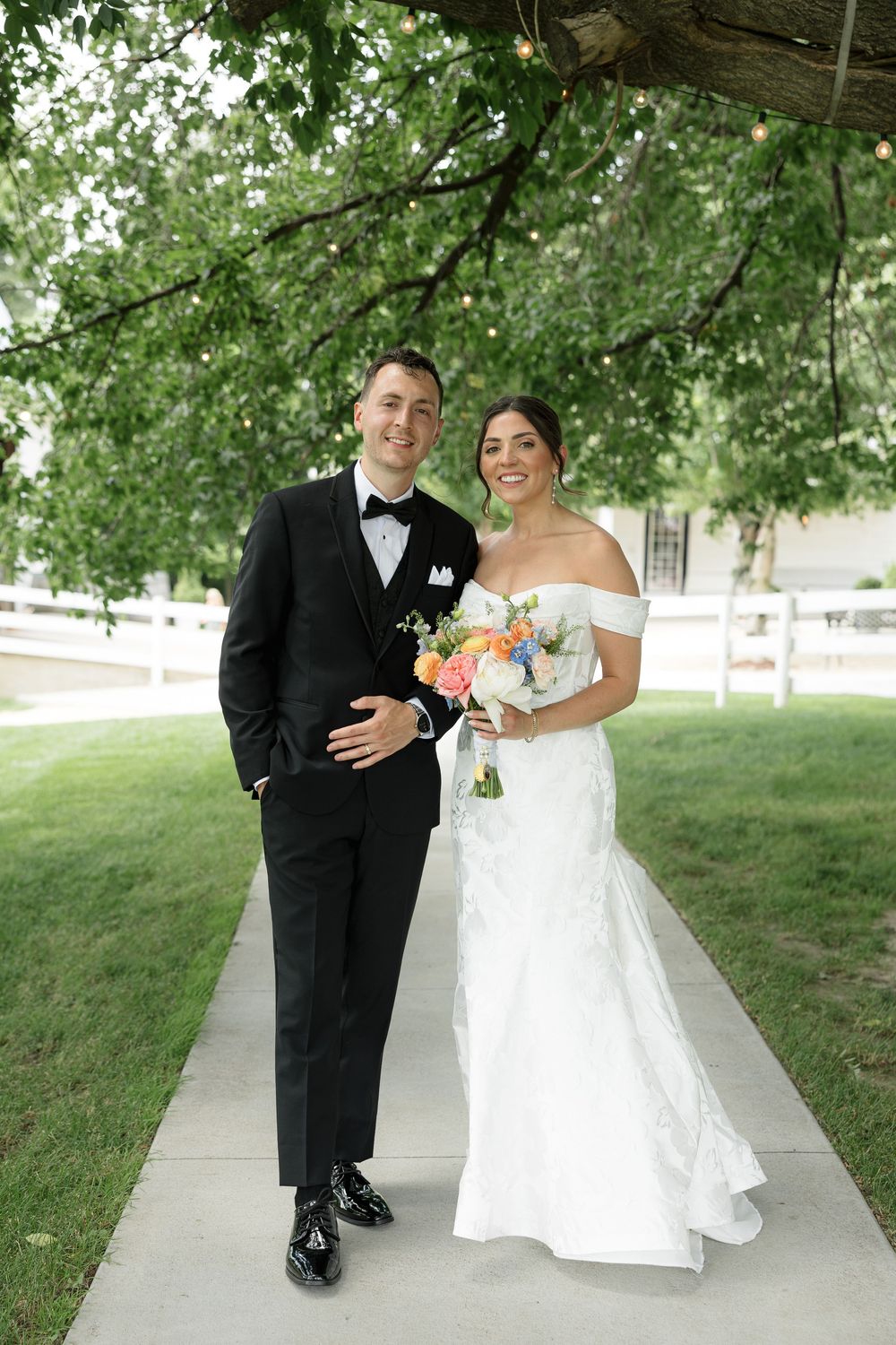 A bride in a white off-shoulder gown and groom in black tuxedo pose together on a pathway under green trees.