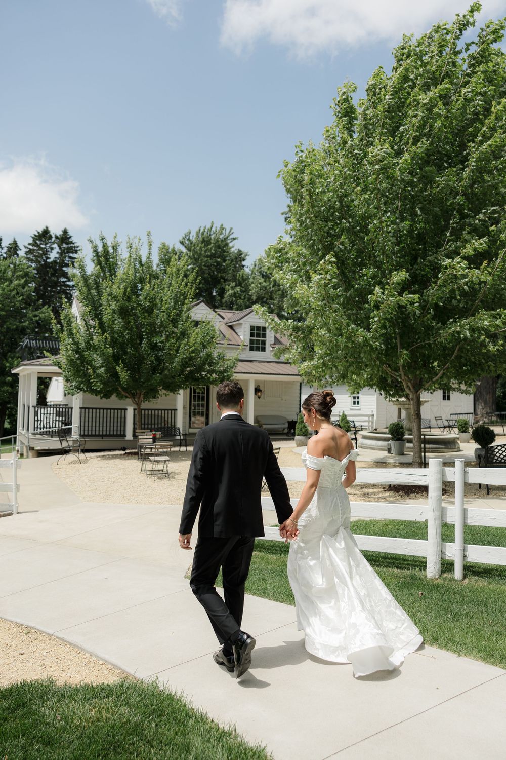 A couple in formal wedding attire walks along a concrete path beside a white fence and green trees on a sunny day.
