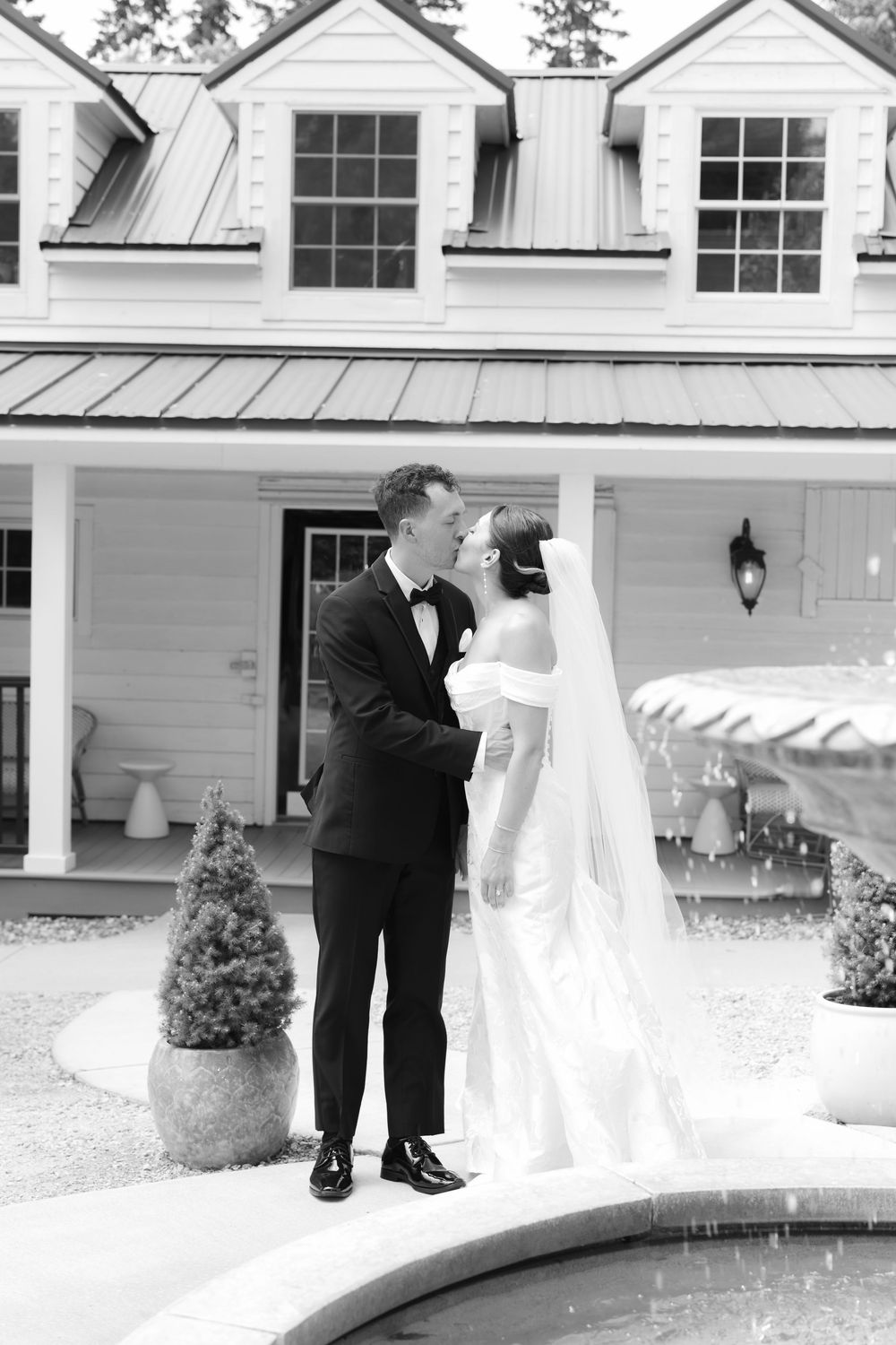 A black and white wedding photo showing a couple in formalwear embracing by a pool in front of a white house with dormers.
