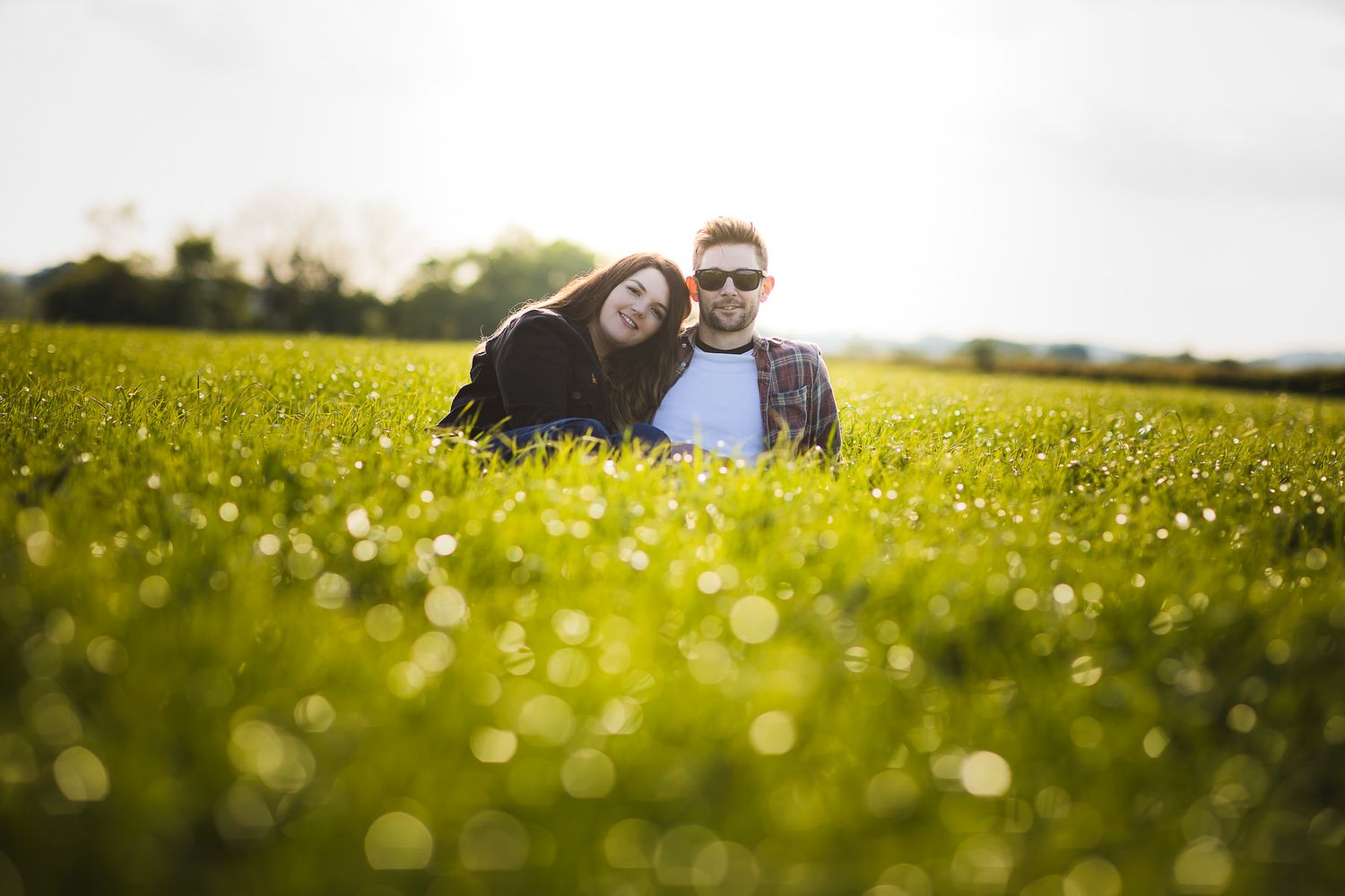 Couple lying in a sunny meadow filled with buttercups as golden light creates bokeh effects in the foreground.