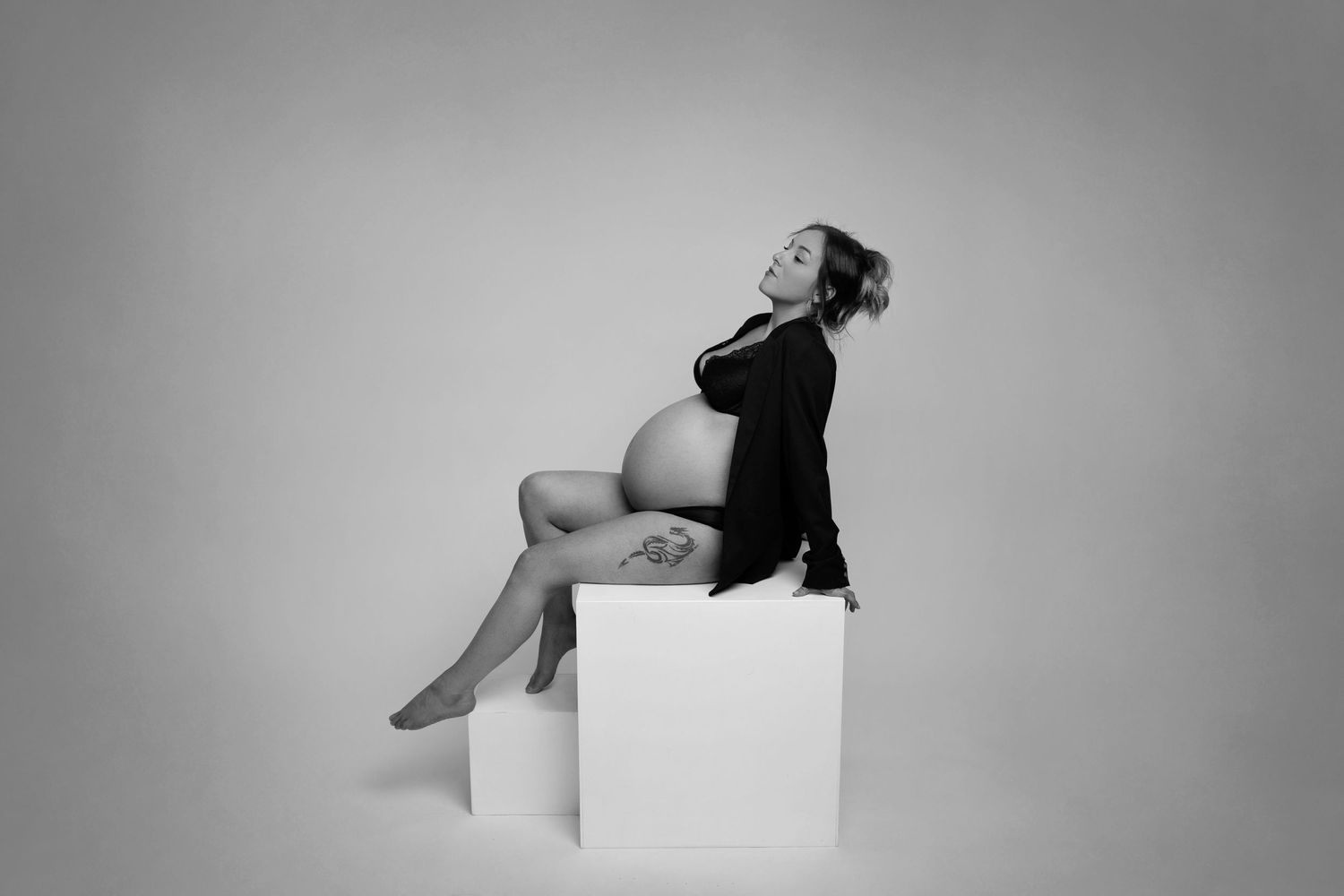 Black and white maternity photo of a woman posing elegantly on a white cube.