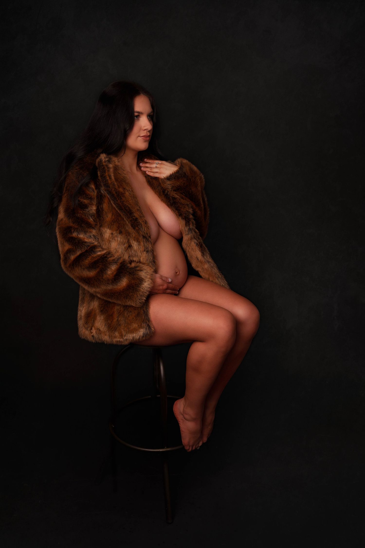 Artistic maternity portrait featuring a woman in a brown fur coat seated on a dark stool.
