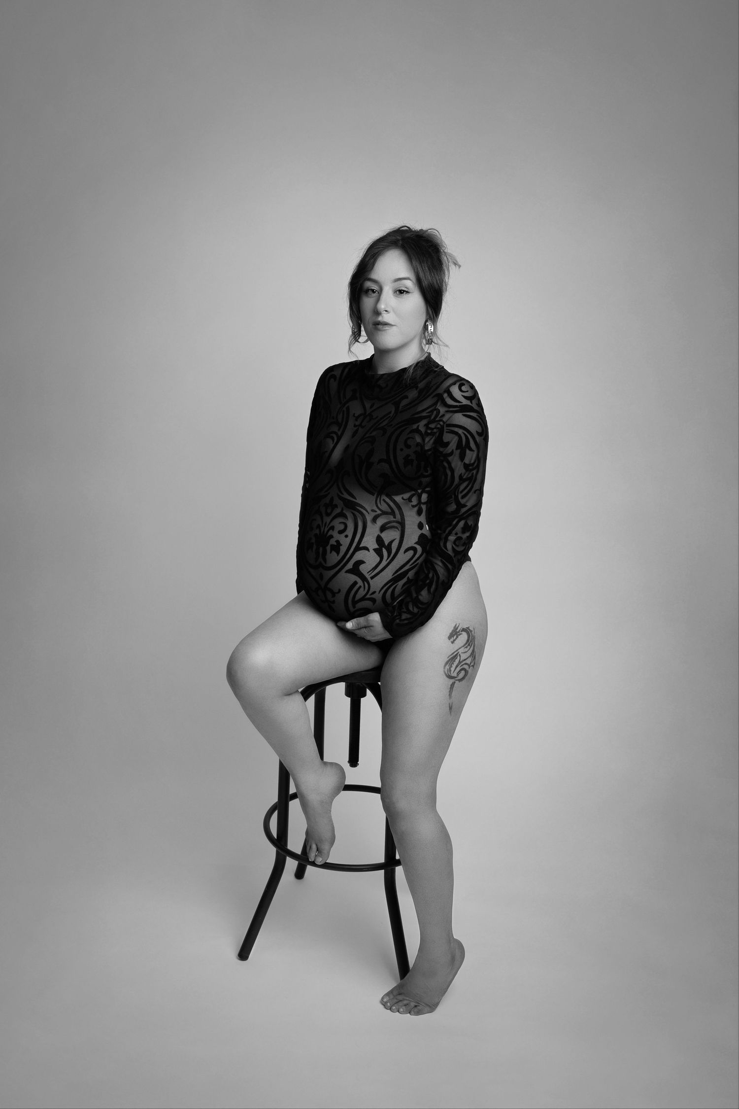 Black and white portrait of a woman in lace dress sitting on stool against light background.