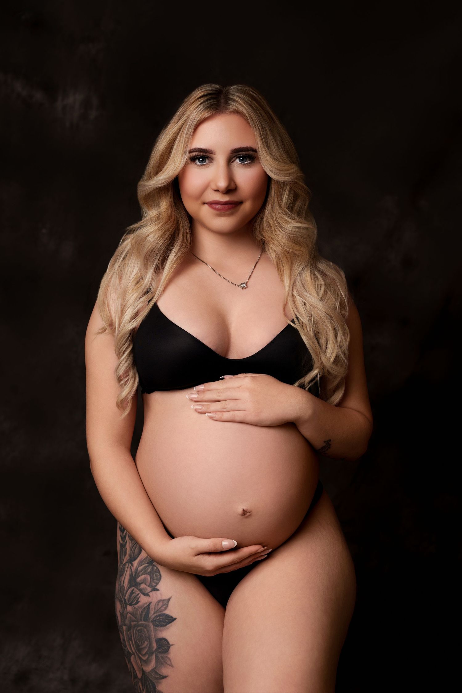 Maternity portrait in black lingerie showing pregnant belly and floral tattoo against dark backdrop.