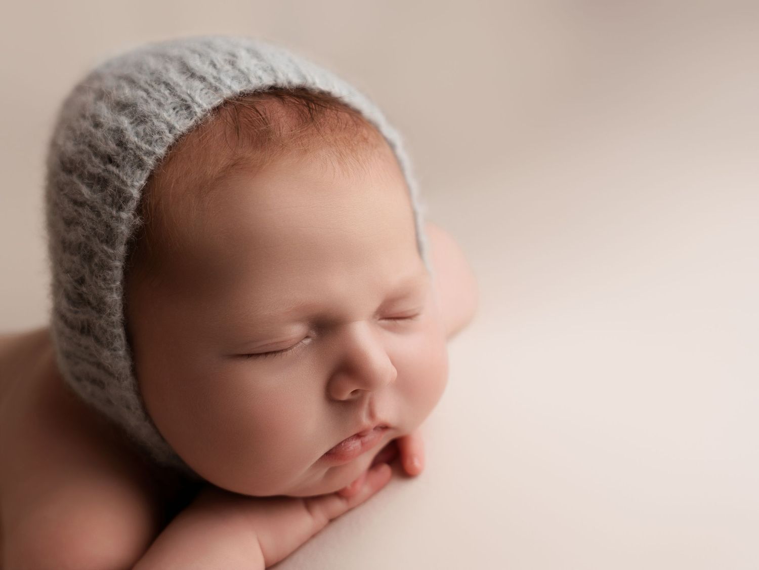 A peaceful sleeping baby wearing a soft gray knit bonnet in a gentle pink-toned newborn portrait.