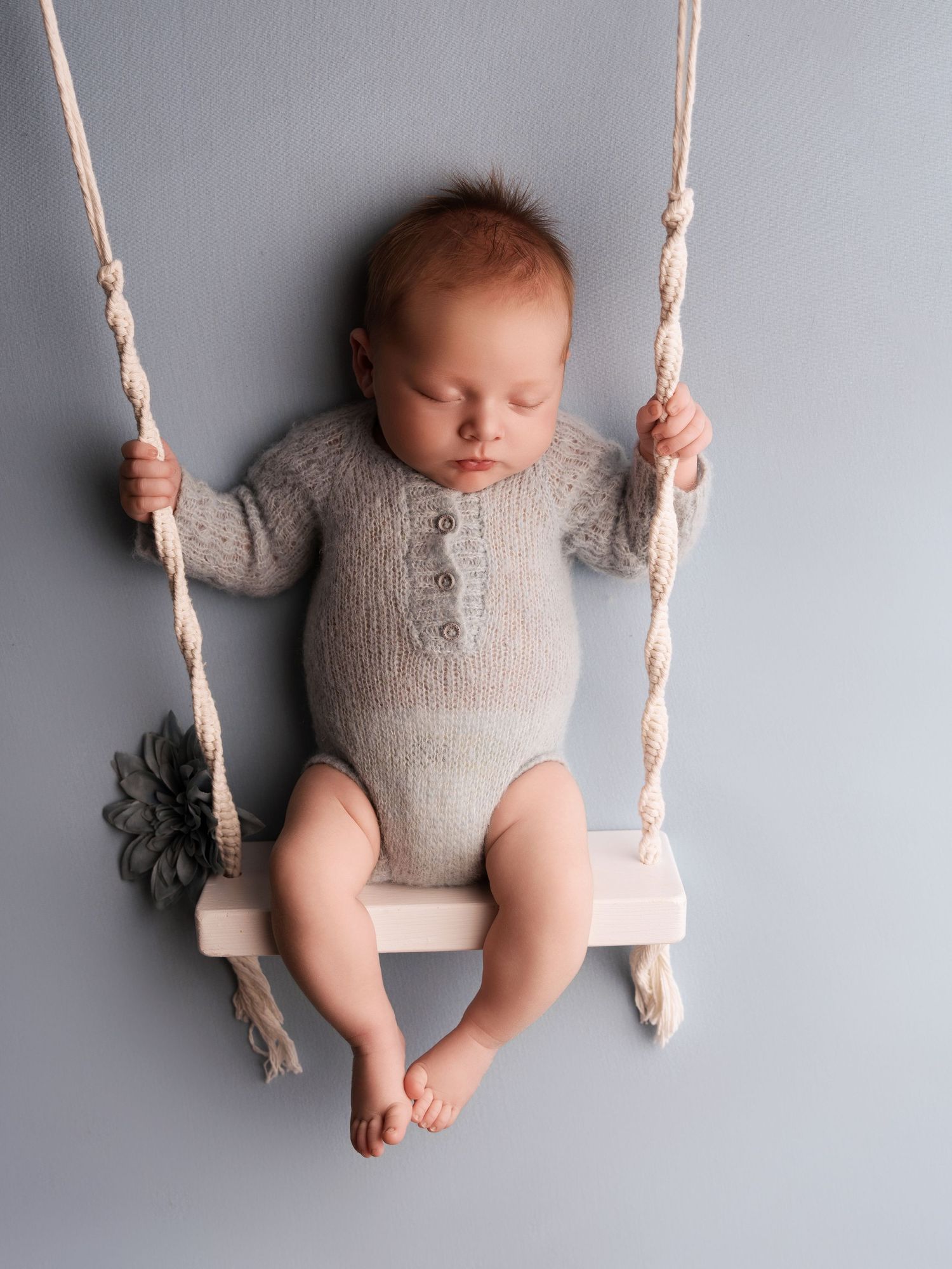 Sleeping baby wearing gray knit romper rests peacefully on small wooden swing with rope handles against light gray backdrop.