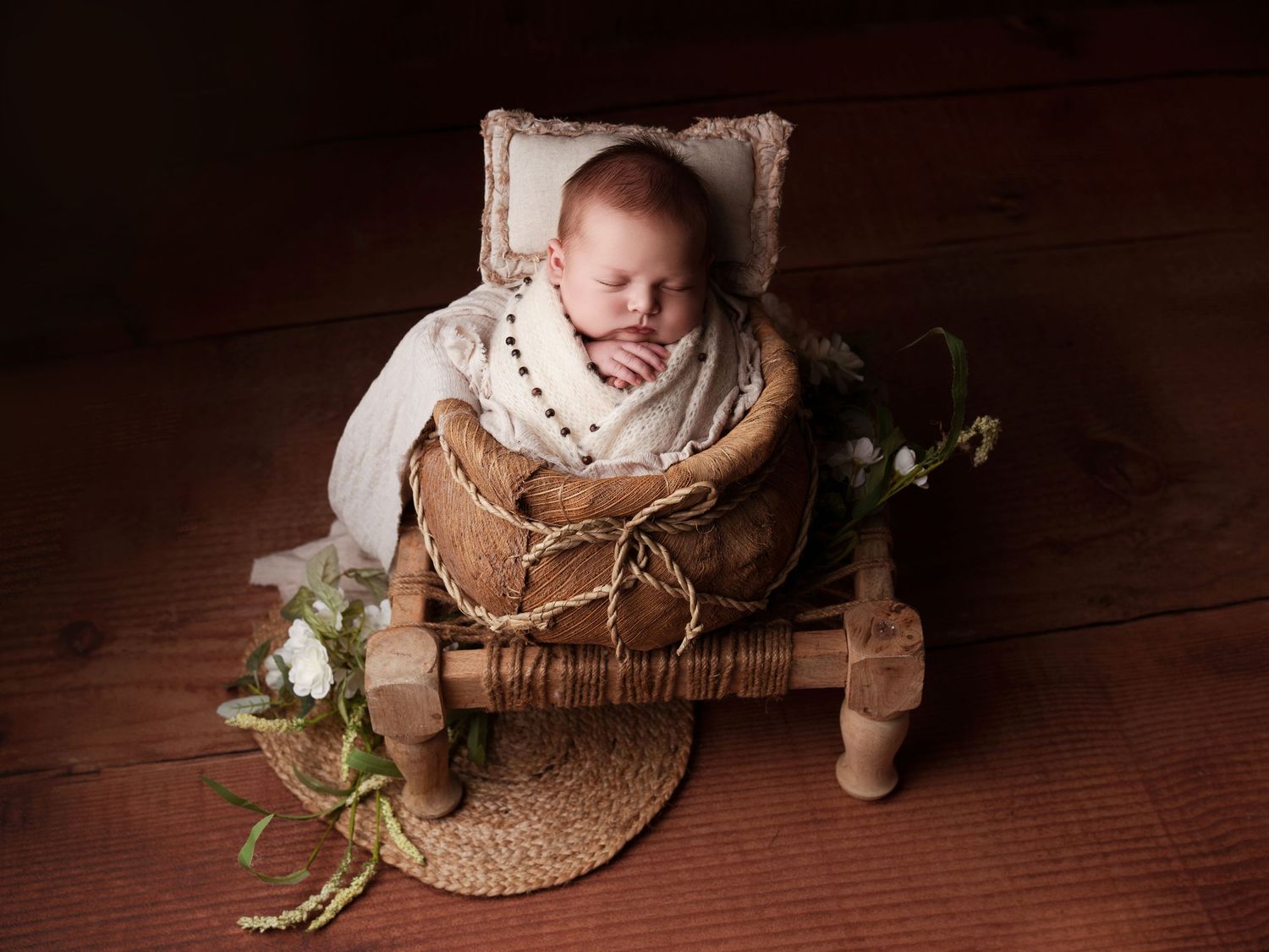 Newborn baby nestled in rustic basket on wooden floor with white swaddle and decorative greenery props for photography session.