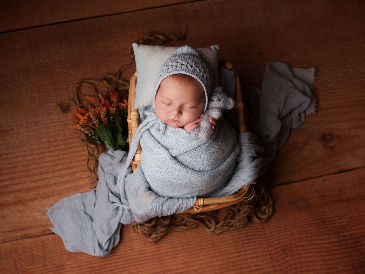 Peaceful sleeping newborn wrapped in soft gray blanket nestled in rustic wooden basket with orange flowers.