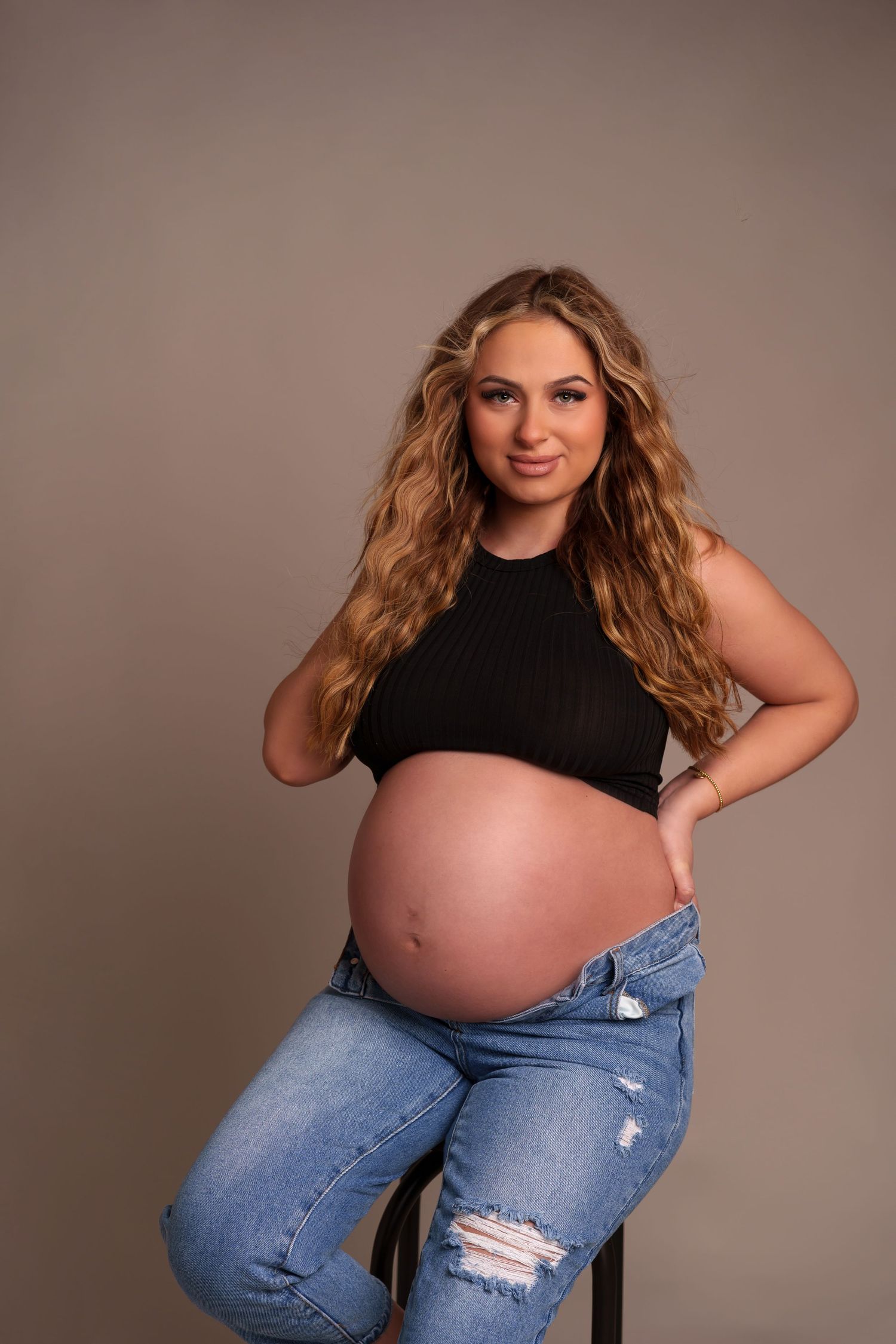 Casual maternity portrait in black top and distressed jeans against neutral background.