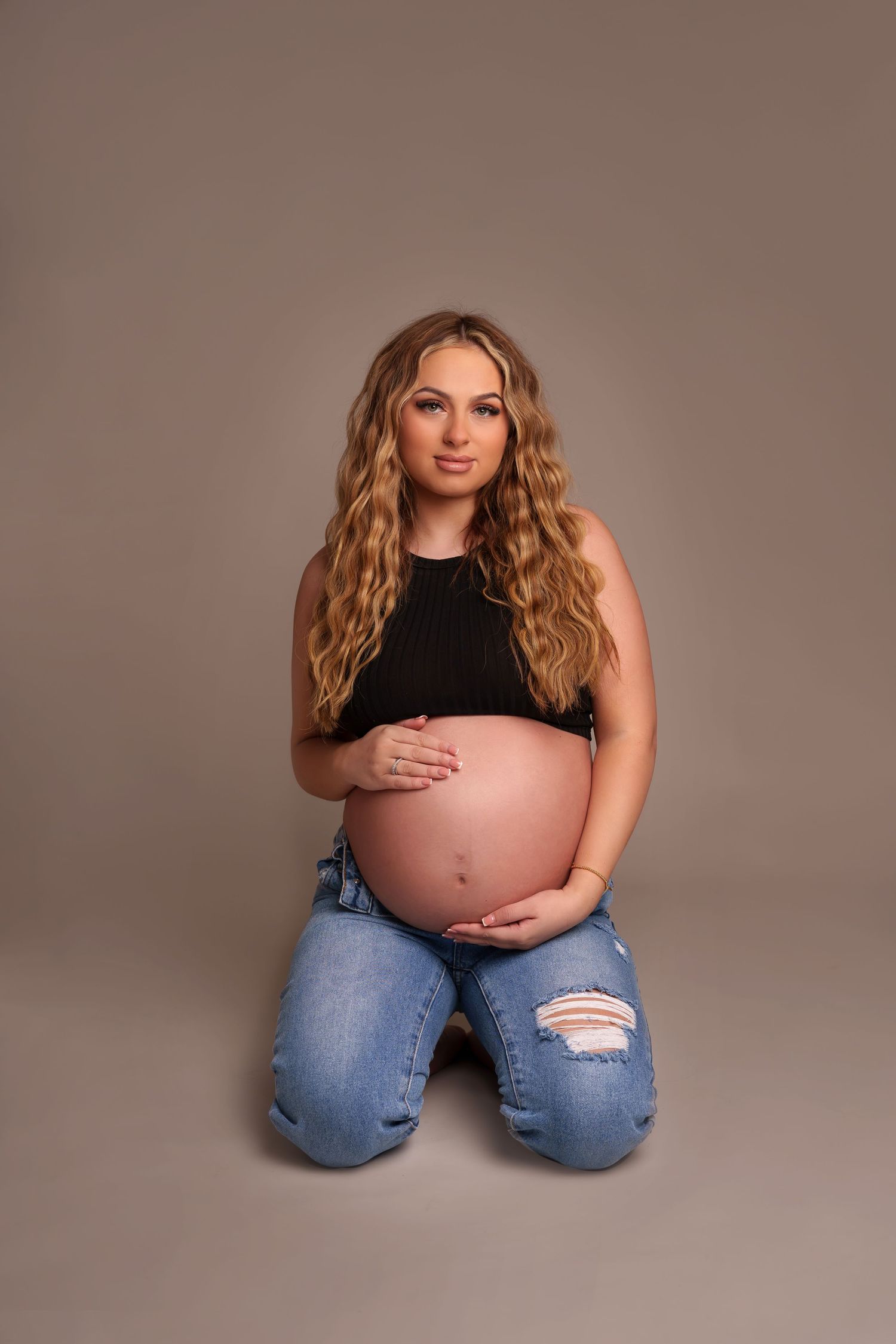 Intimate maternity pose in black tank top and ripped jeans on neutral backdrop.