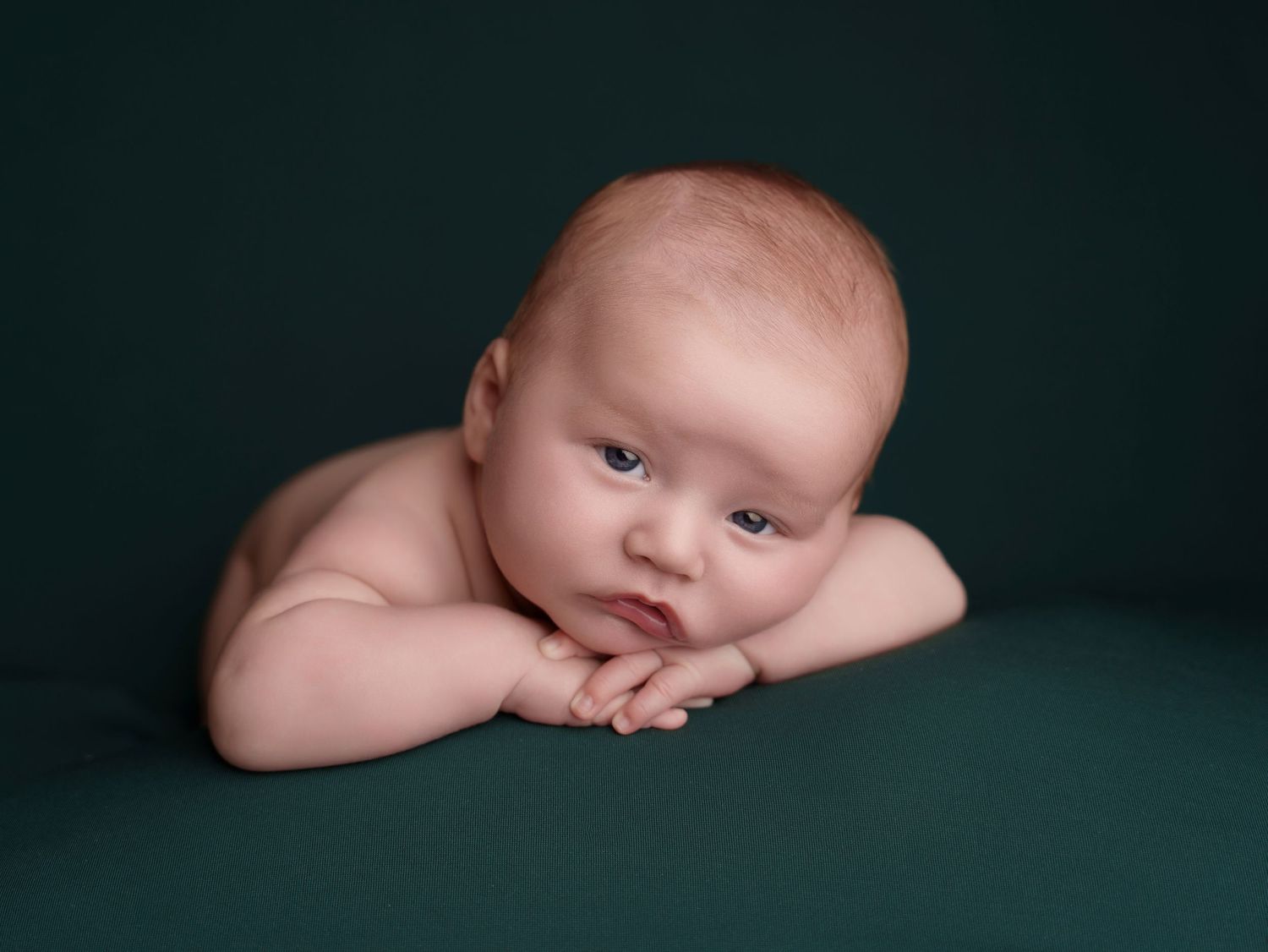 Sweet newborn baby resting with folded arms on dark green background during professional portrait session.
