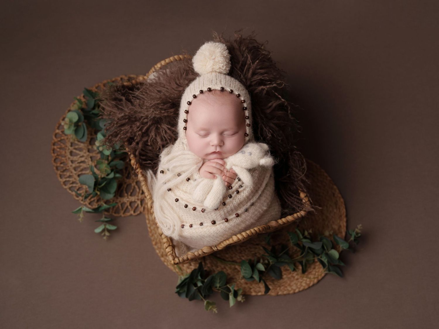 Sleeping newborn swaddled in white knit wear rests peacefully in woven basket with green ivy decorative accents.