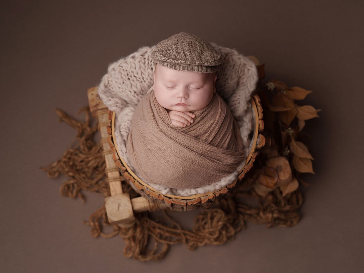 A sleeping newborn wrapped in beige fabric lies in a wooden bowl surrounded by dried autumn leaves.