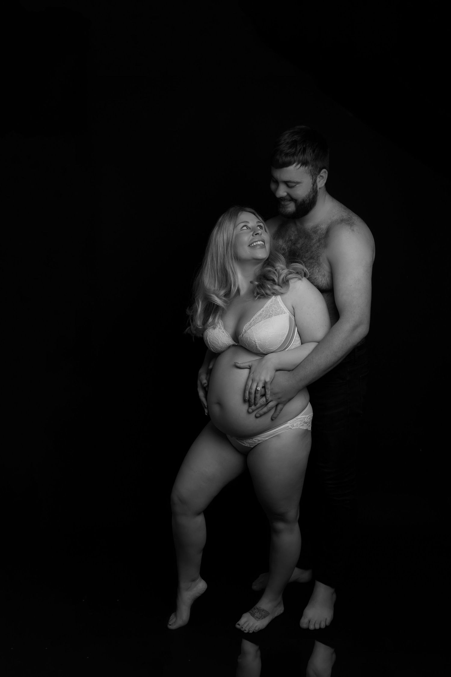 Intimate black and white maternity photo of an expectant couple embracing in a dark studio setting.