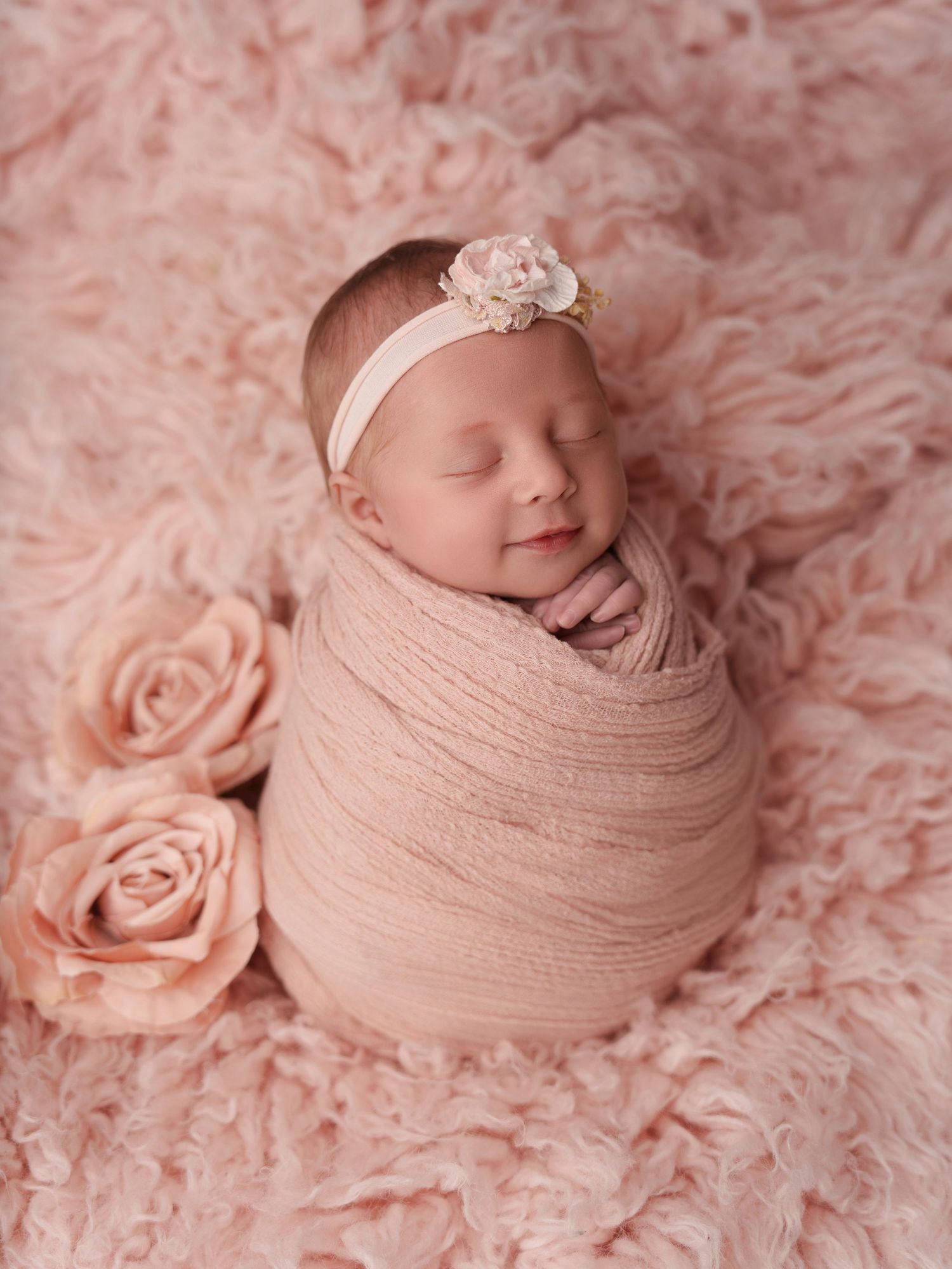 Sleeping newborn wrapped in soft pink fabric with floral headband on pink rose textured background.