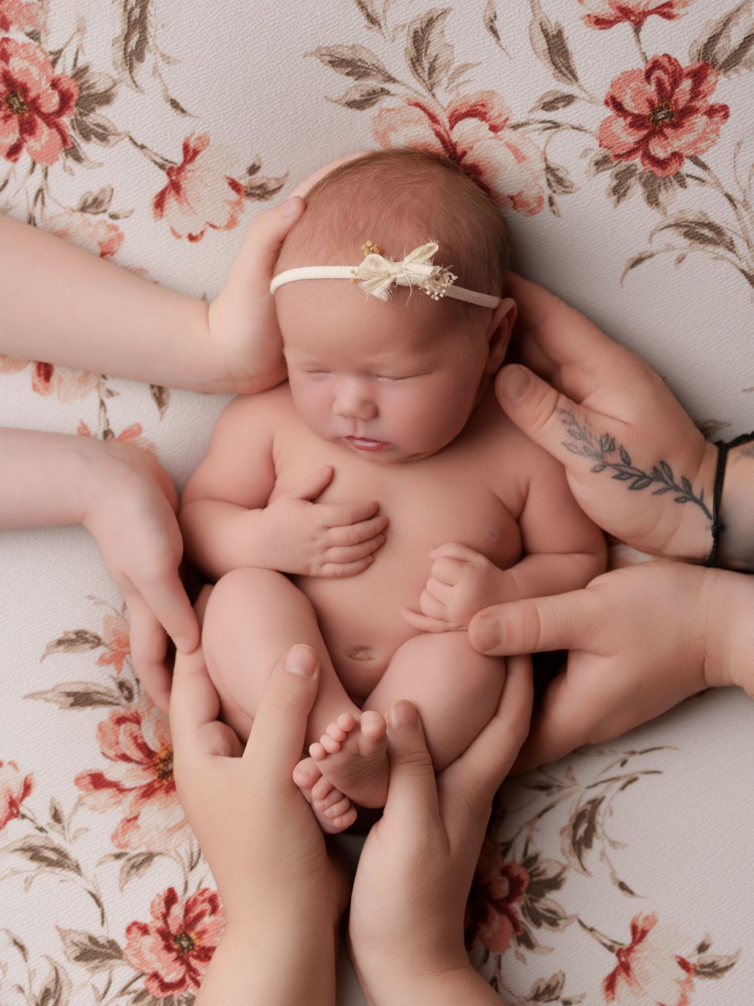 A sleeping newborn in a cream headband lies on a floral background while gentle hands support their tiny body.