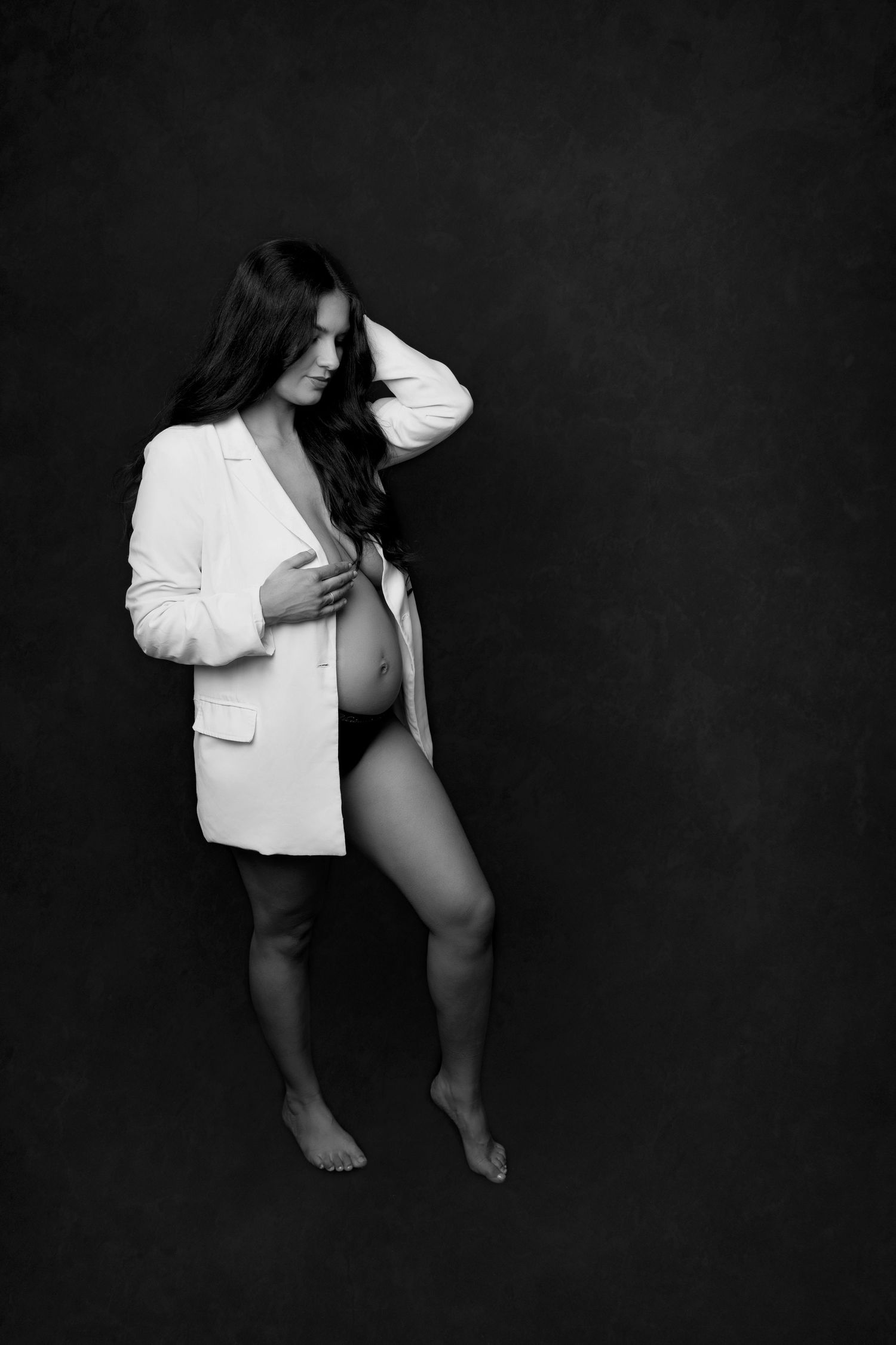 Dramatic black and white portrait of a pregnant woman wearing a white blazer against a dark background.
