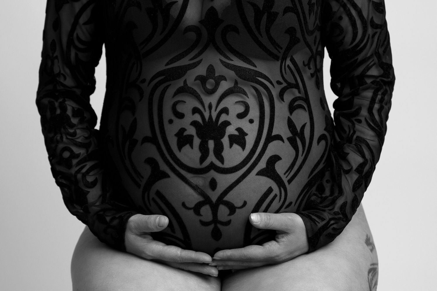 Close-up black and white photo of hands cradling a pregnant belly in a patterned dress.