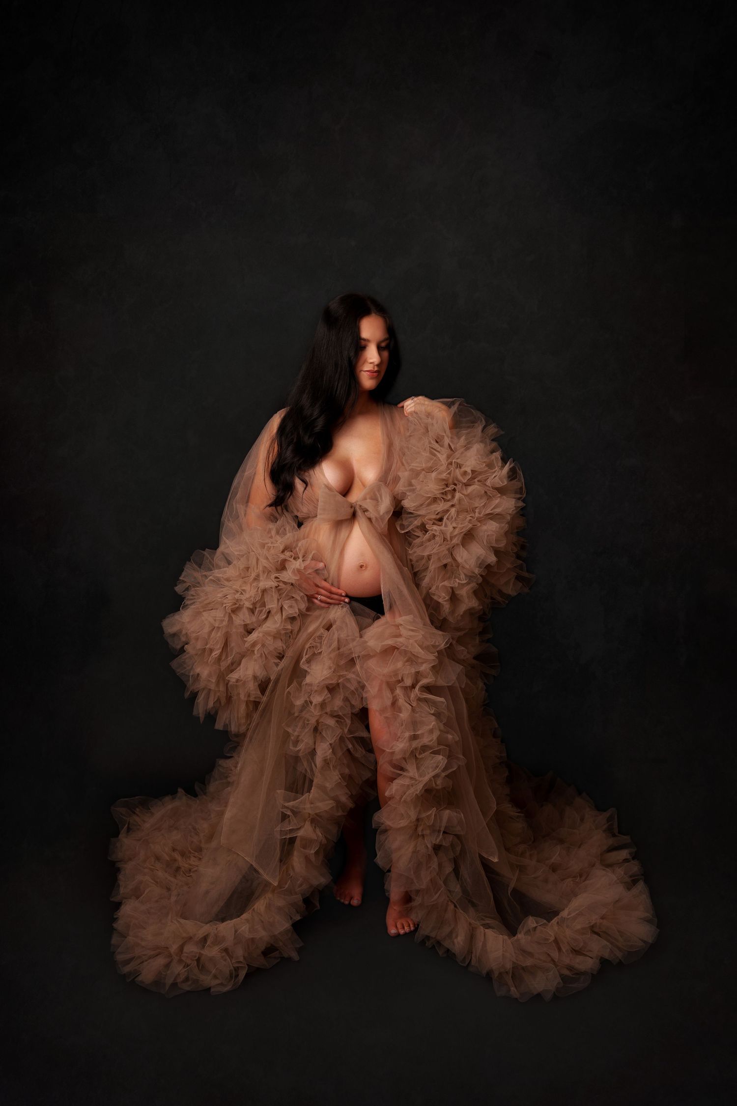 Artistic maternity portrait with a flowing tan tulle gown against a dark backdrop.