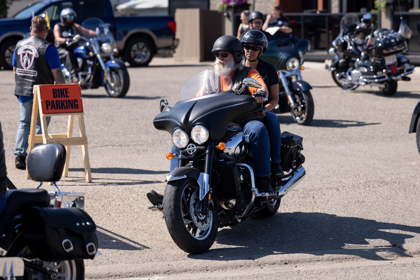 Row of motorcycles lined up at an outdoor event with blue sky and parked vehicles in the background.