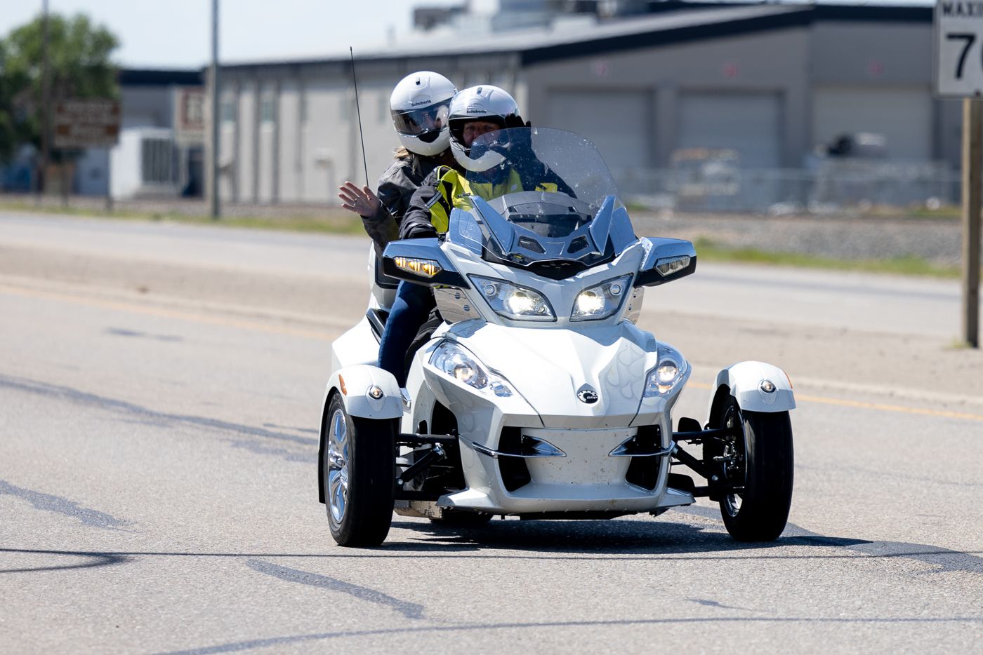 Two riders cruise on a white Can-Am Spyder three-wheeled motorcycle on a sunny street.