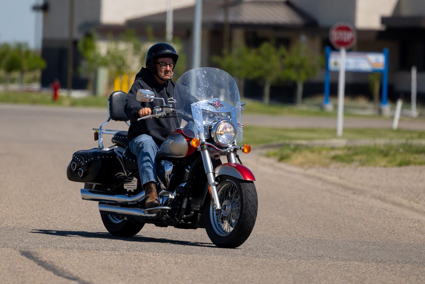 A motorcyclist rides a classic cruiser motorcycle equipped with saddlebags on a sunny suburban street.