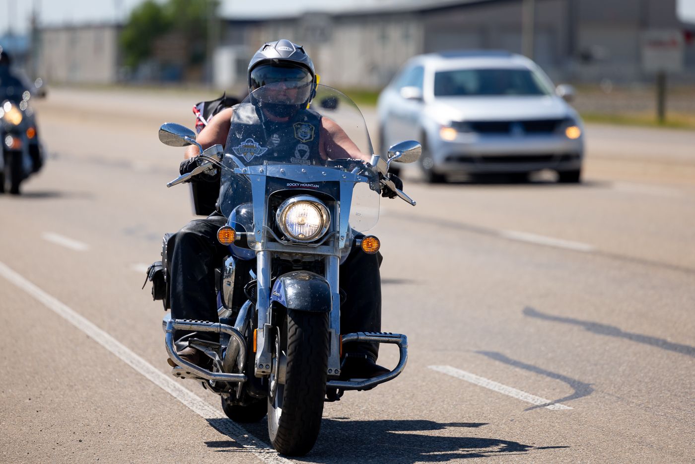 A motorcyclist rides a black cruiser motorcycle on a sunlit road with vehicles in the background.