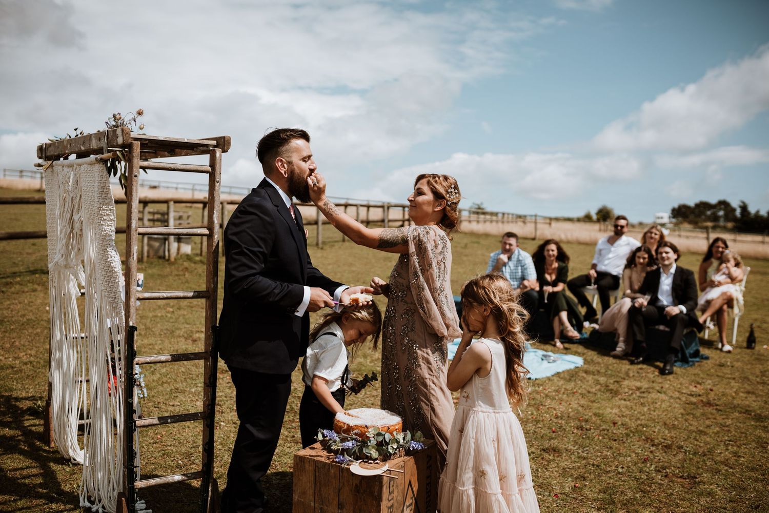 An outdoor countryside wedding ceremony with a rustic wooden arch and guests seated on the hillside under blue skies.