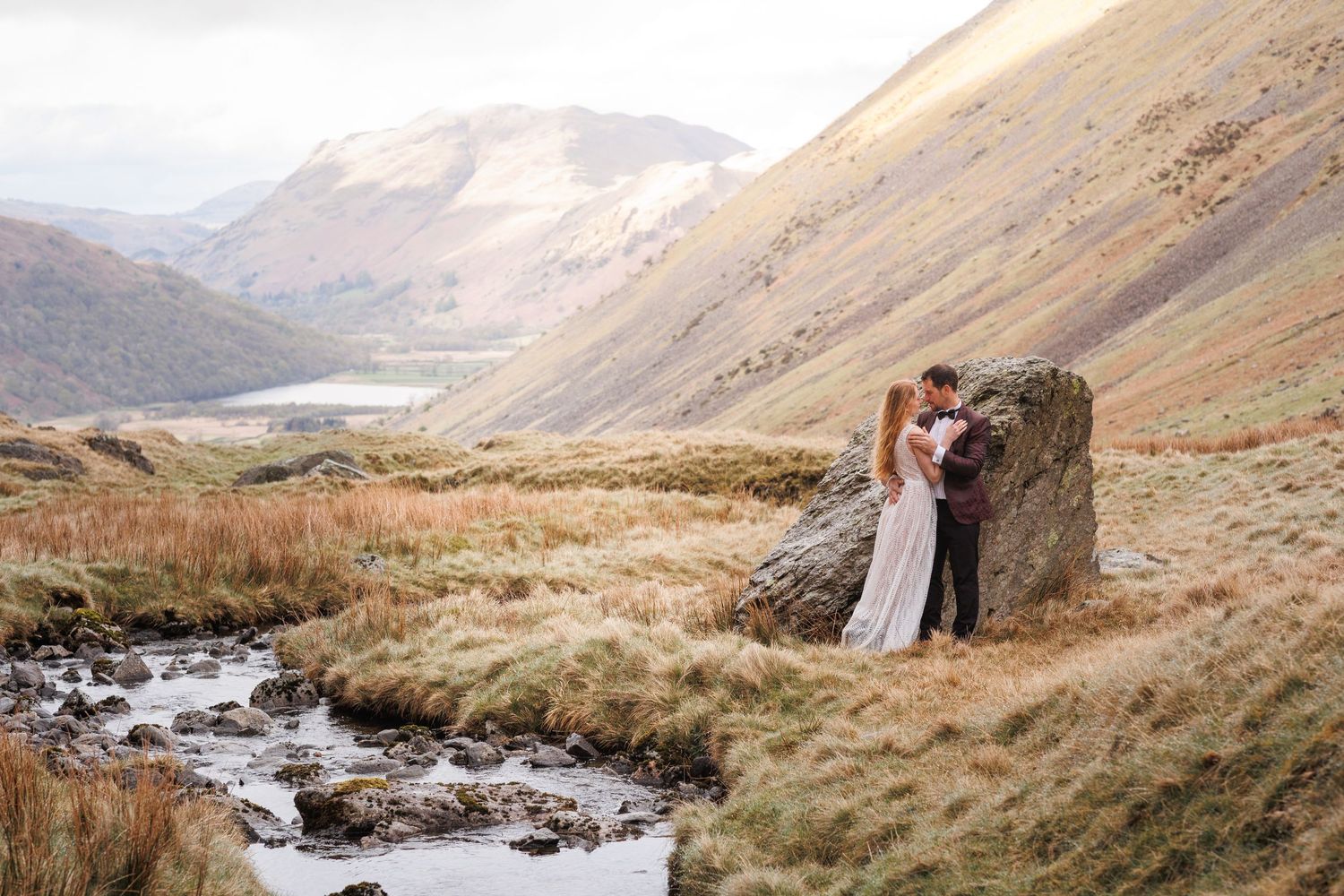 Dramatic mountain landscape with winding stream and rolling hills in muted tones.