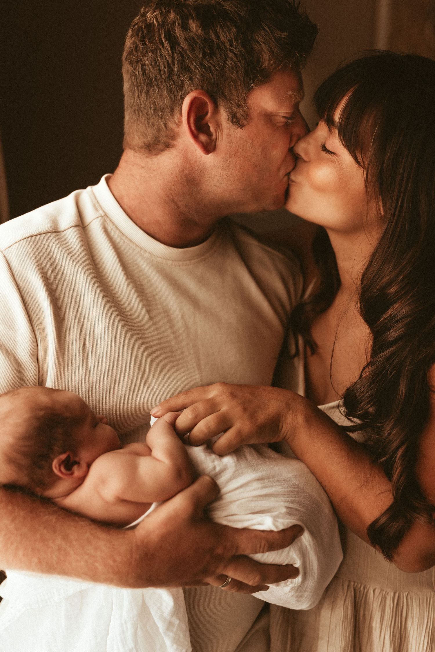 A tender family moment as parents share a kiss while holding their newborn baby in a warm, intimate setting. Newborn photographer Javea